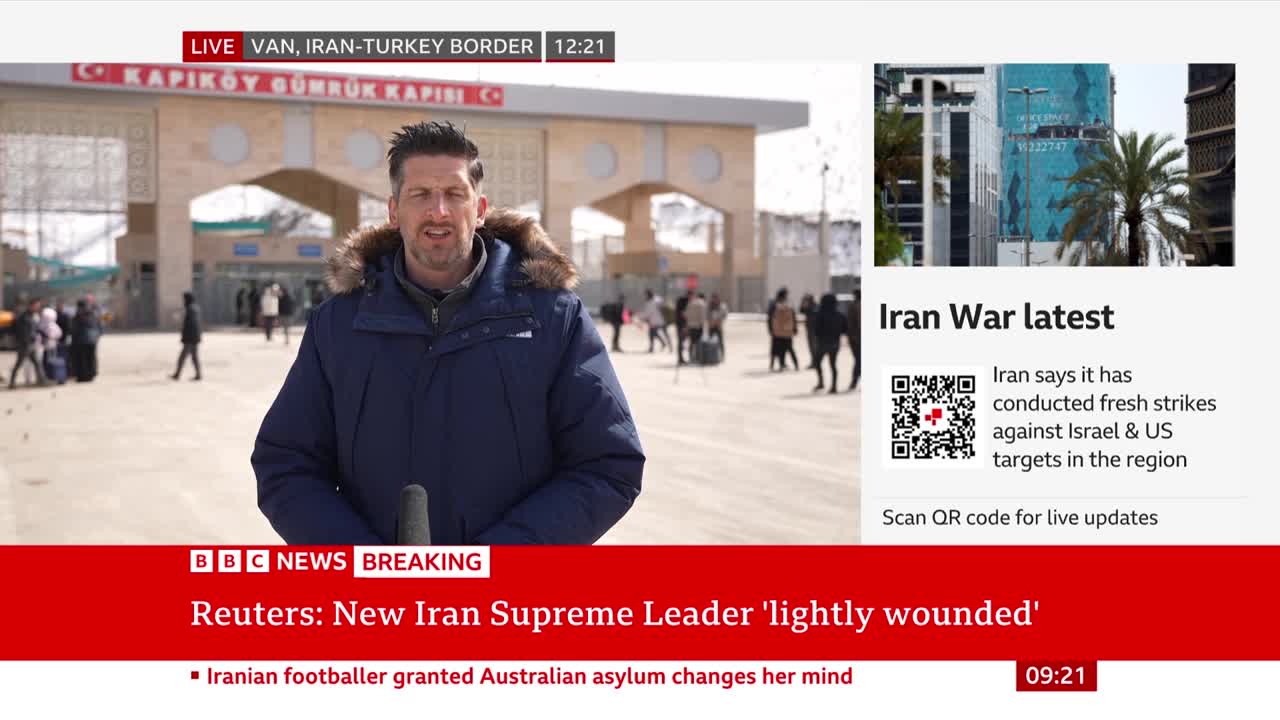 A reporter stands before a border crossing, a microphone in hand. People are moving through the gate behind him, some with luggage. A reporter stands before a border crossing, a microphone in hand. People are moving through the gate behind him, some with luggage.