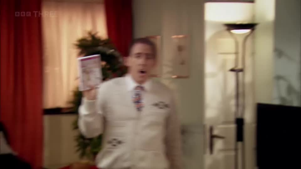 A man in a white cardigan and patterned tie yelps, holding up a DVD. The BBC Three logo is visible in the corner. A man in a white cardigan and patterned tie yelps, holding up a DVD. The BBC Three logo is visible in the corner.