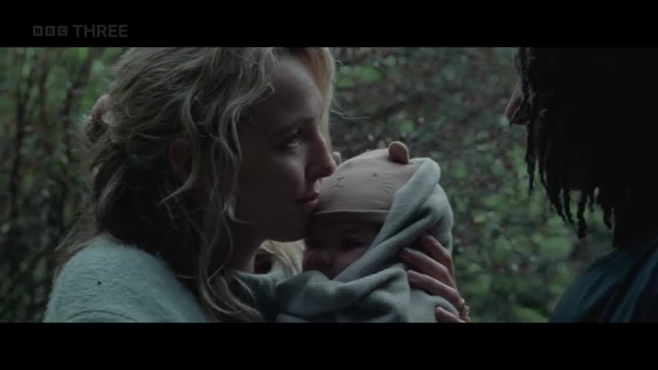 A woman gently kisses the forehead of a swaddled baby. Beside them, a man with dreadlocks watches them, his face mostly in shadow. A woman gently kisses the forehead of a swaddled baby. Beside them, a man with dreadlocks watches them, his face mostly in shadow.