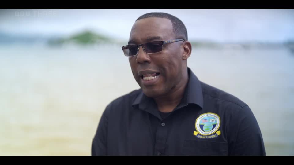 A man in a black polo shirt with sunglasses is speaking, a BBC Three logo visible in the corner. The background is a hazy expanse of water. A man in a black polo shirt with sunglasses is speaking, a BBC Three logo visible in the corner. The background is a hazy expanse of water.