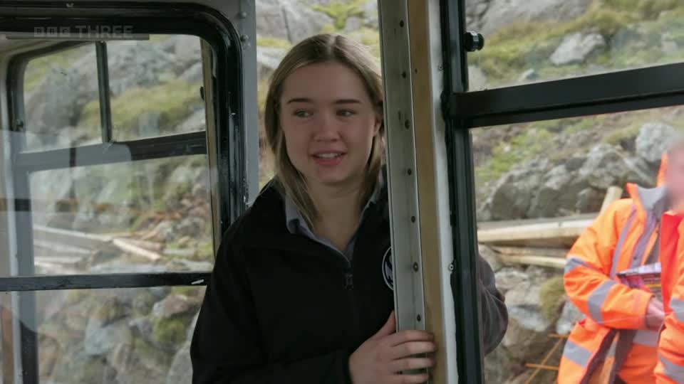 A young woman in a black jacket stands inside a tram, looking out at the rocky, mountainous landscape. Two men in bright orange high-visibility jackets are visible through the tram's windows. A young woman in a black jacket stands inside a tram, looking out at the rocky, mountainous landscape. Two men in bright orange high-visibility jackets are visible through the tram's windows.