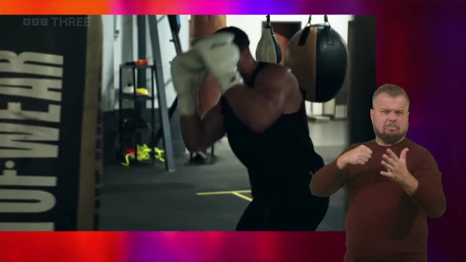 A boxer, wearing white gloves, throws a punch at a speed bag. Beside him, a man signs with his hands, his expression earnest. A boxer, wearing white gloves, throws a punch at a speed bag. Beside him, a man signs with his hands, his expression earnest.