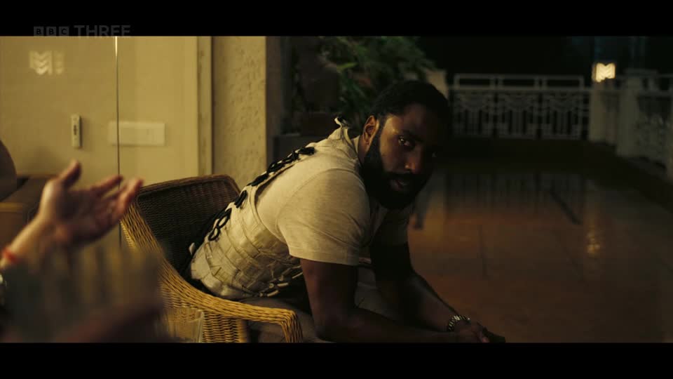 The man in the tan vest leans forward, his gaze intense. A hand gestures to his left, its fingers splayed.