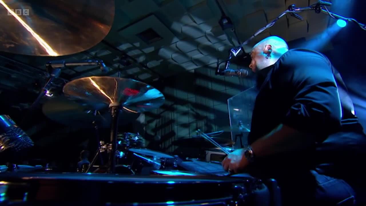The drummer's sticks are a blur as he hits the snare, a bright blue light illuminating his focused expression. This performance is being broadcast live on BBC Two.