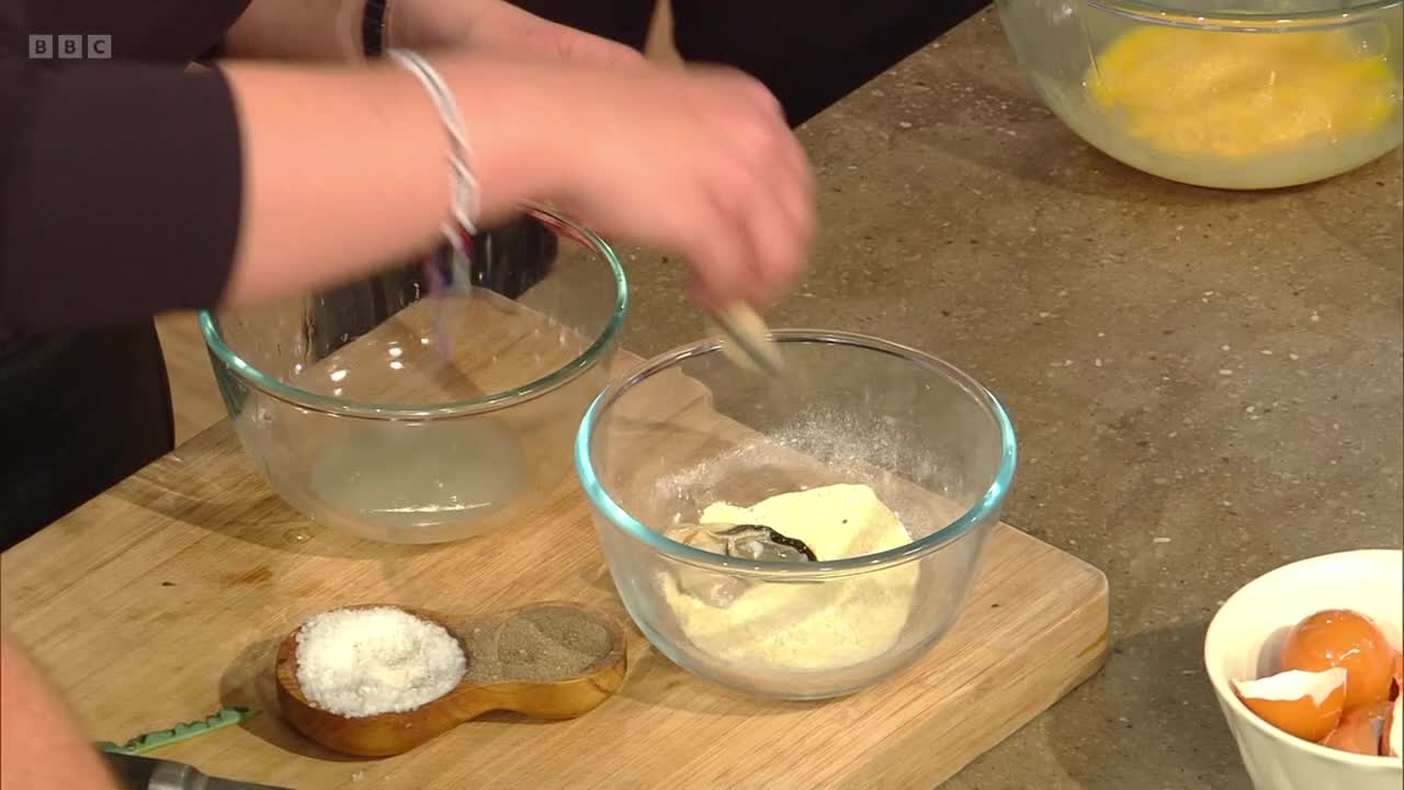 A hand dips something into a bowl of flour, then moves towards another bowl. A wooden spoon holds salt and pepper nearby.
