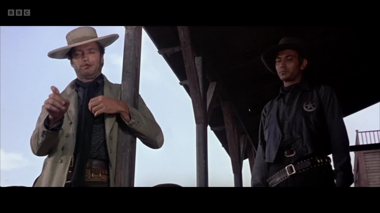 A man in a wide-brimmed hat gestures with his left hand, his right adjusting his dark scarf. Another man, wearing a sheriff's badge, stands stoically to his right. The BBC Two logo is visible in the corner of the screen.