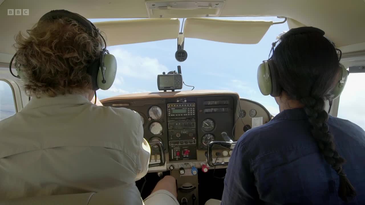 The pilot's hands are on the controls of the small plane. Beside them, a passenger watches the sky through the cockpit window.