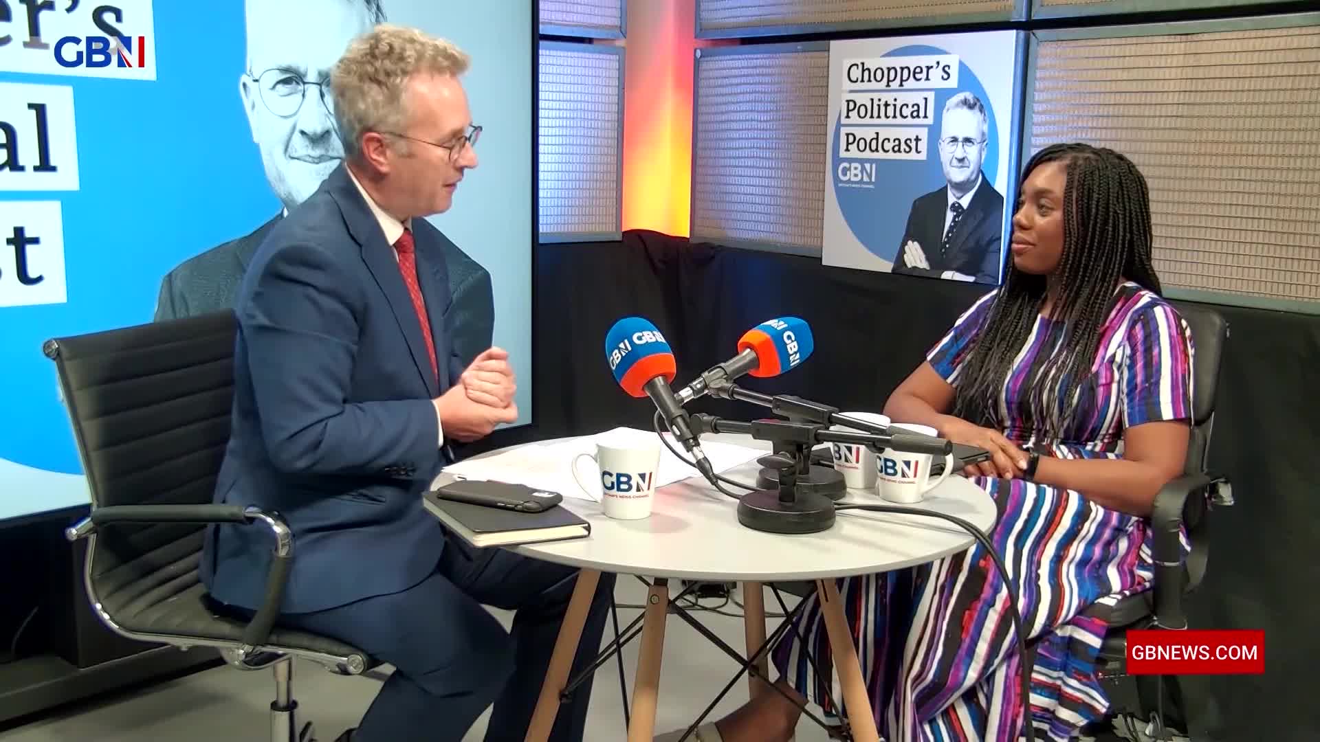 A man in a suit and tie speaks into a microphone, facing a woman with braided hair. They are seated at a round table with two GB News branded mugs and more microphones.