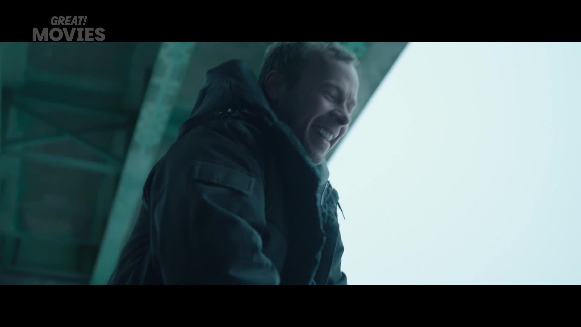 A man in a dark jacket laughs heartily, his face crinkled with amusement. He's standing under a bridge, the pale sky behind him.