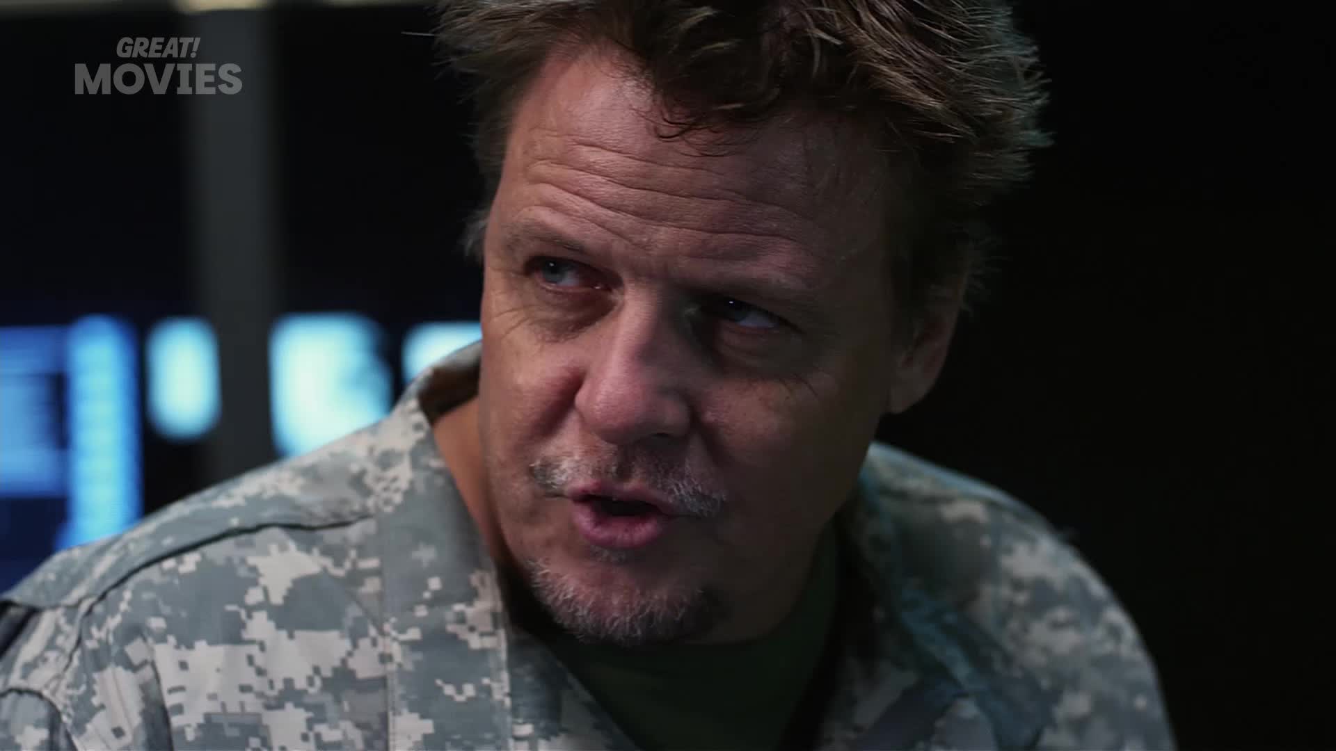 A man in a camouflage uniform speaks intently. He has a weathered face and a greying mustache.