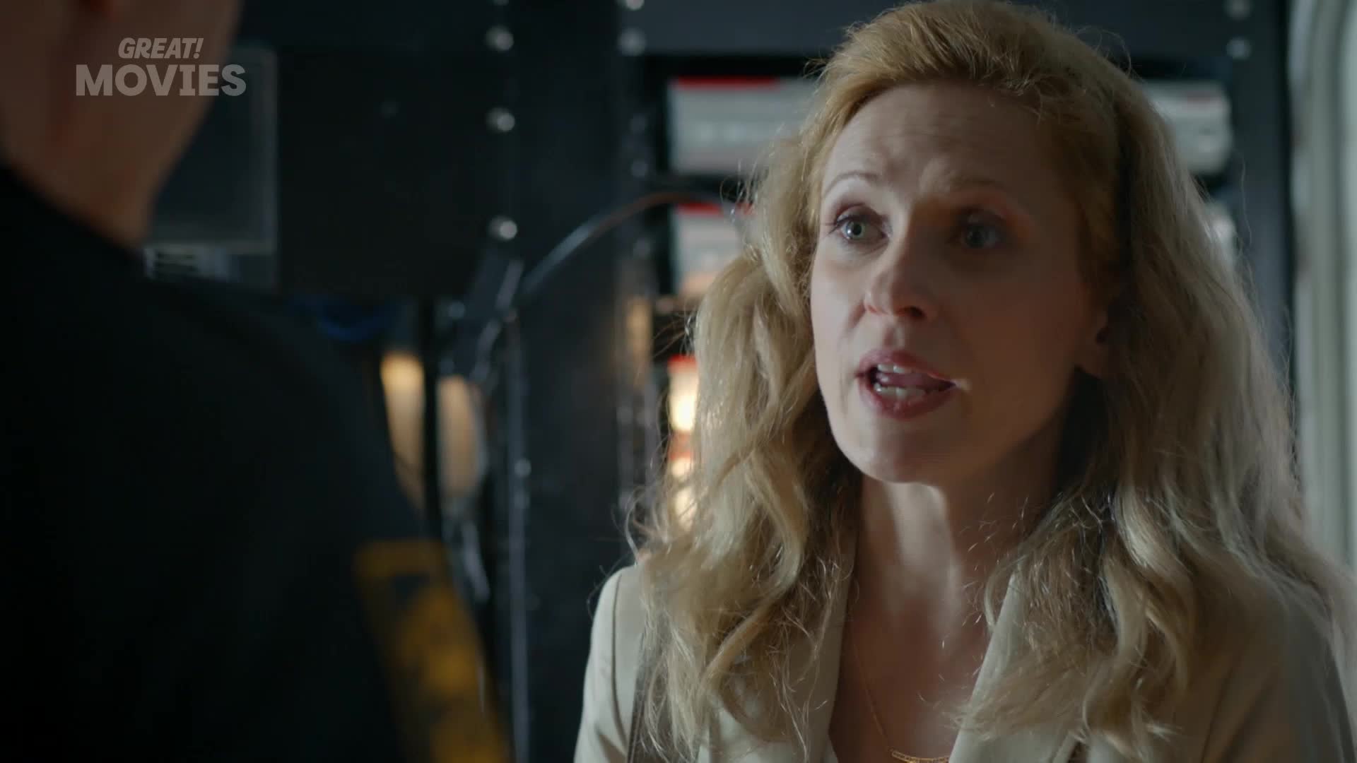 A woman with blonde, wavy hair speaks intently, her mouth slightly open. Behind her, a man in a dark uniform is partially visible, his shoulder blurred from movement.