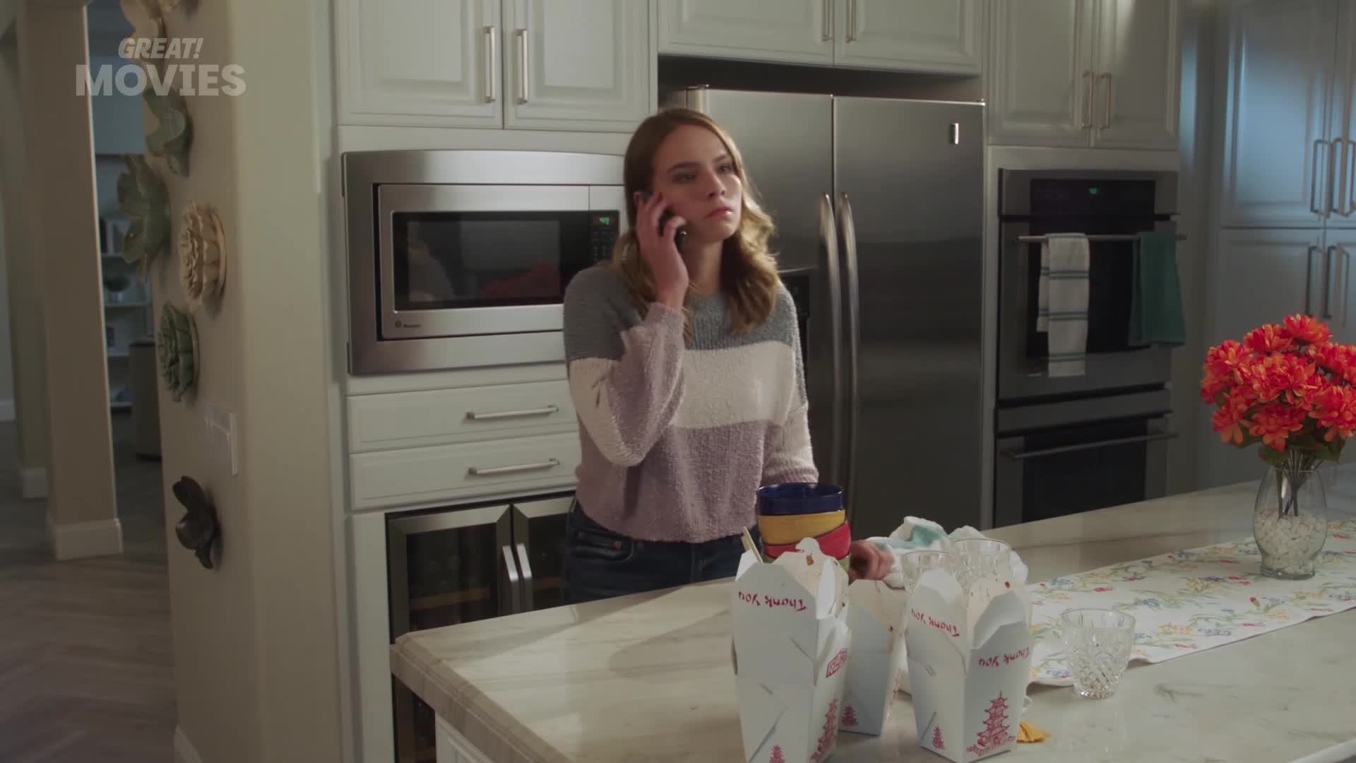 A young woman stands by the counter, talking on her phone, with several takeaway containers lined up. A vase of bright orange flowers sits on the counter to her right.