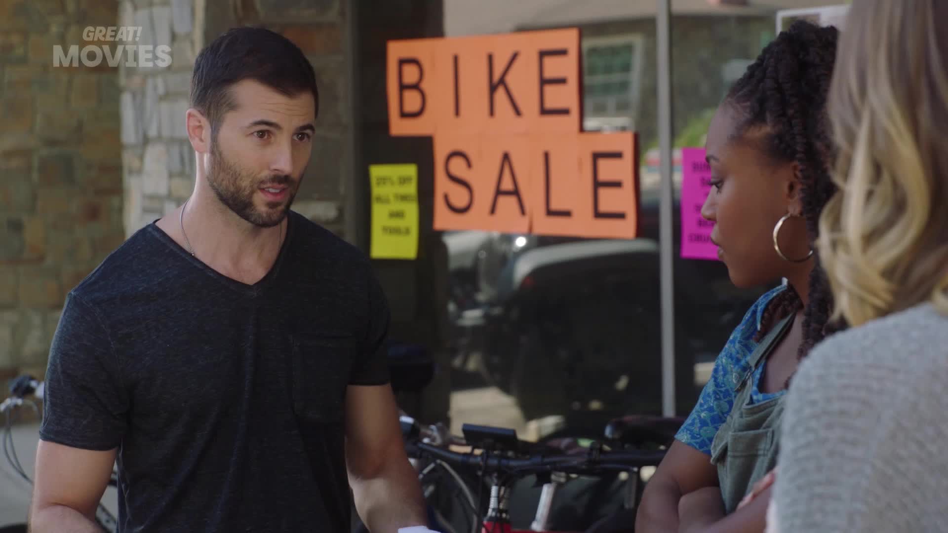 A man in a dark t-shirt gestures as he speaks to two women outside a shop with a "BIKE SALE" sign in the window. The women, one with large hoop earrings, listen intently.
