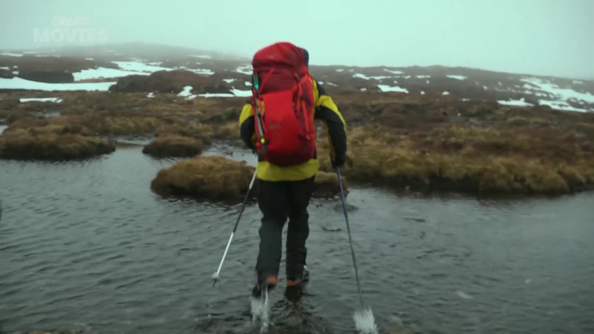 A lone hiker, clad in yellow and black with a bright red pack, navigates a shallow, rocky stream. They use trekking poles to steady themselves on the uneven ground, pushing through the water in a misty, moorland landscape.