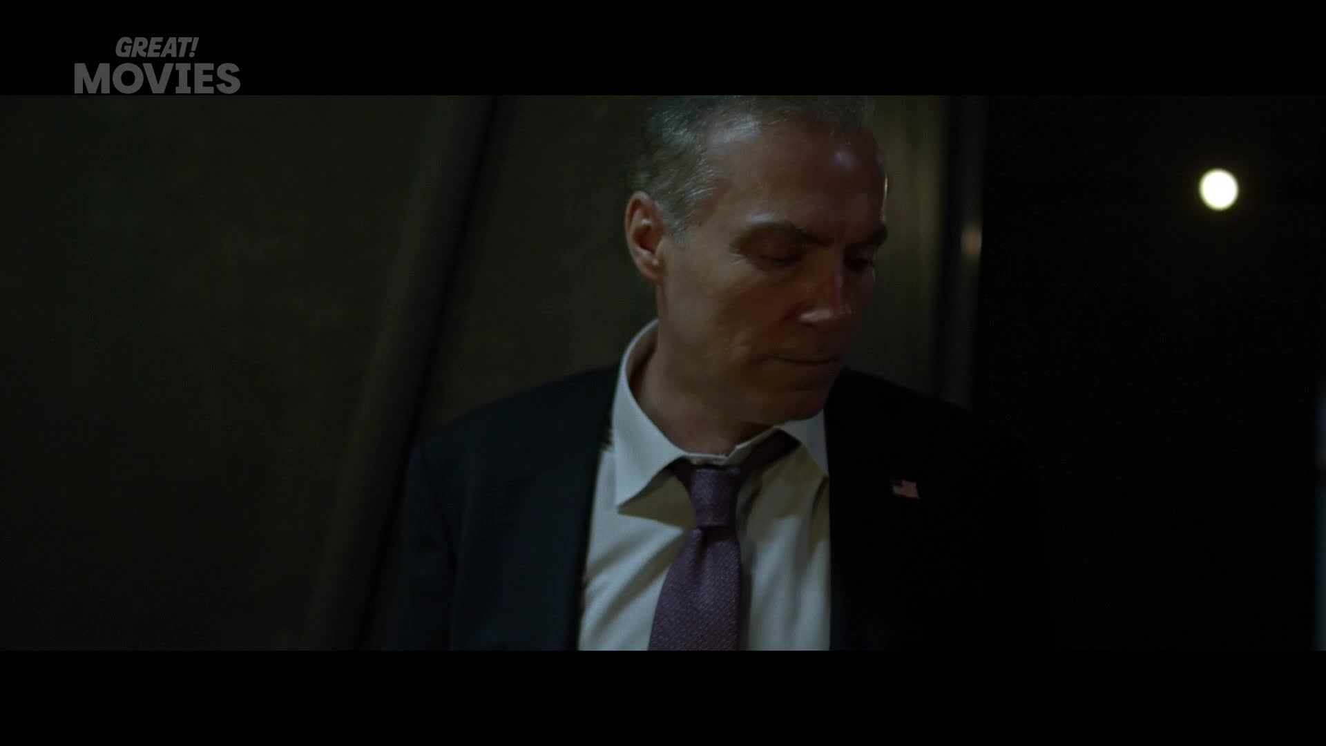 A man in a dark suit and tie looks down, his face shadowed. A faint light gleams in the background, suggesting a late hour in a city somewhere in the UK.