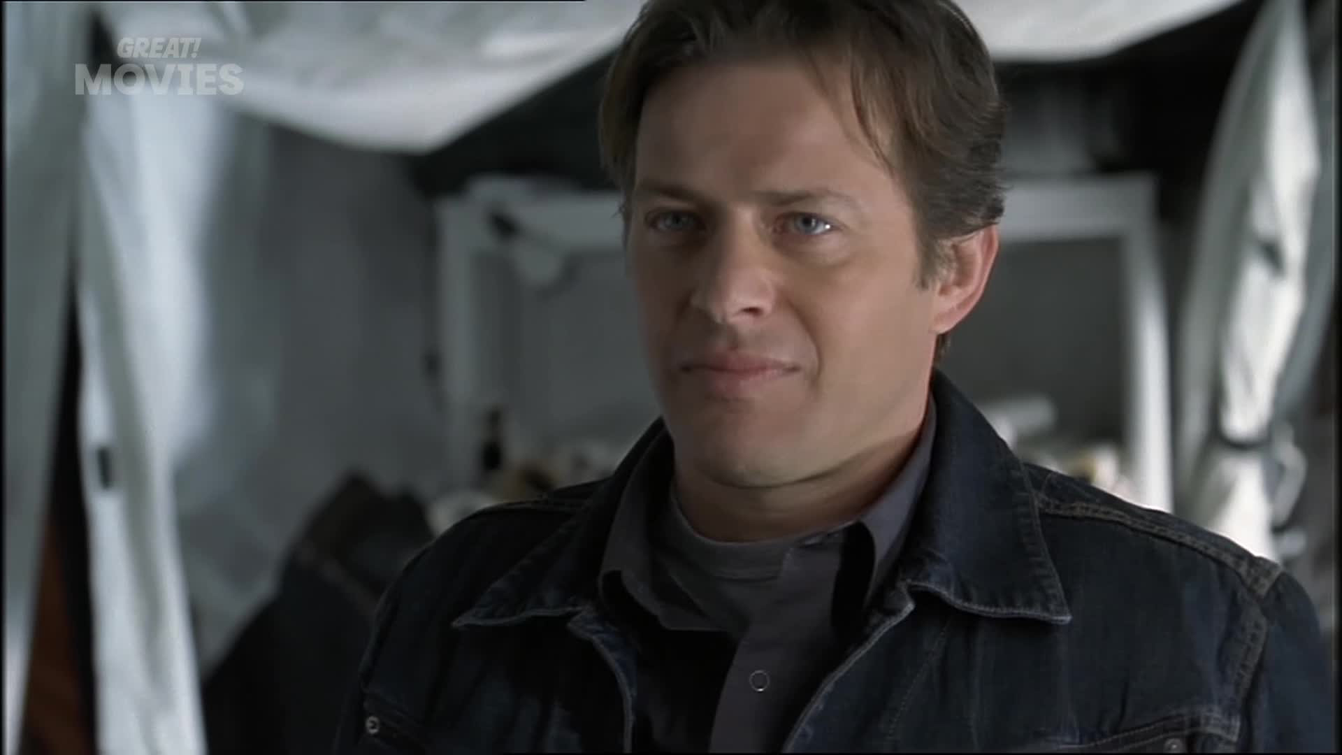 A man in a denim jacket looks to his right with a slight smirk. The background is out of focus, with white sheeting and some shelving visible.