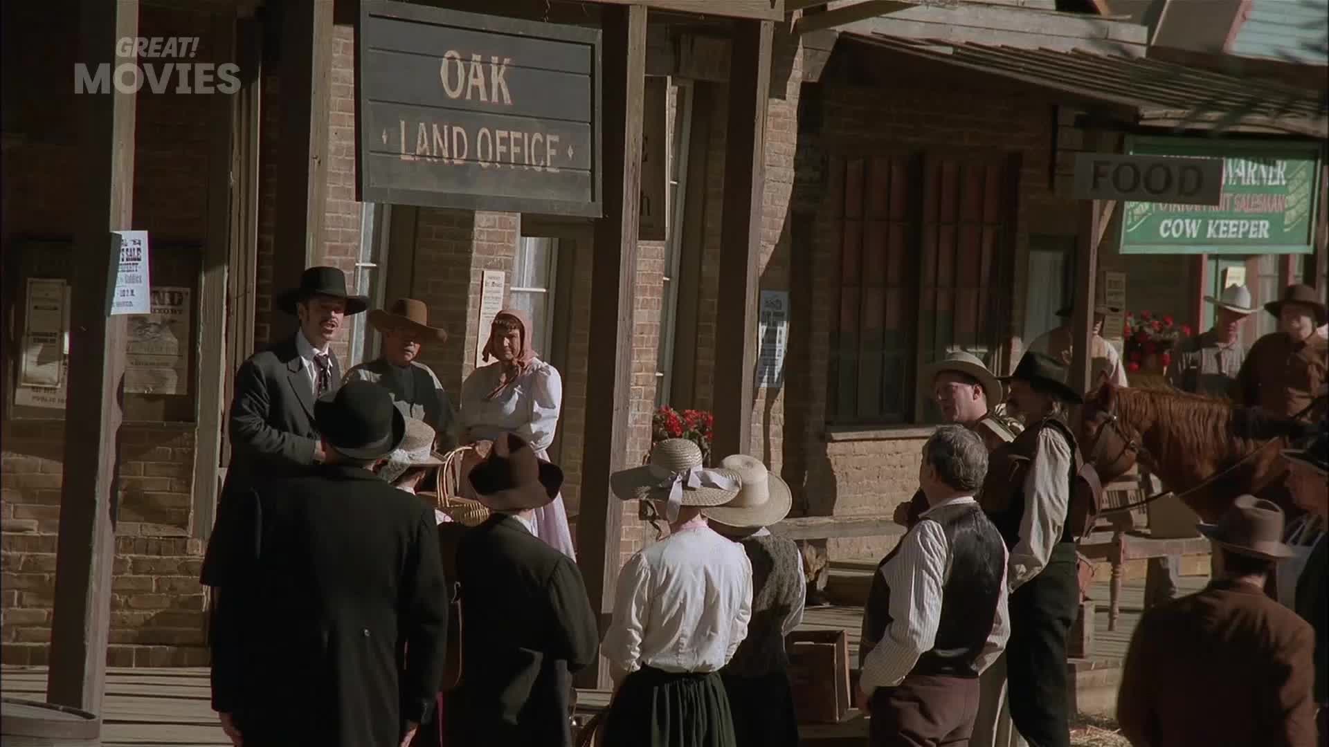 A man in a suit and hat speaks with a woman in a white dress, while others gather outside the Oak Land Office. A horse-drawn cart passes by the food market, its driver tipping his hat.