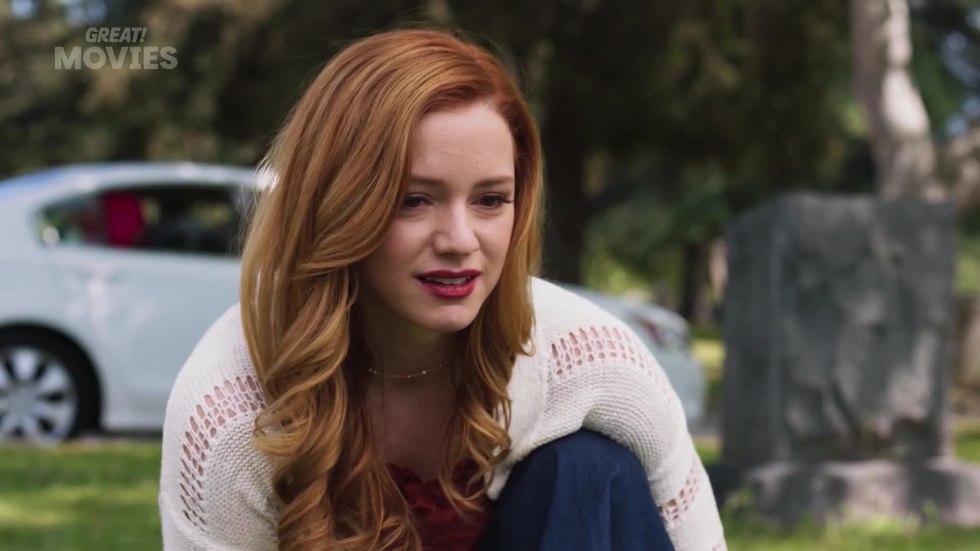 A young woman with bright auburn hair kneels in a grassy area. A white car is parked behind her, and a weathered stone marker stands in the background.