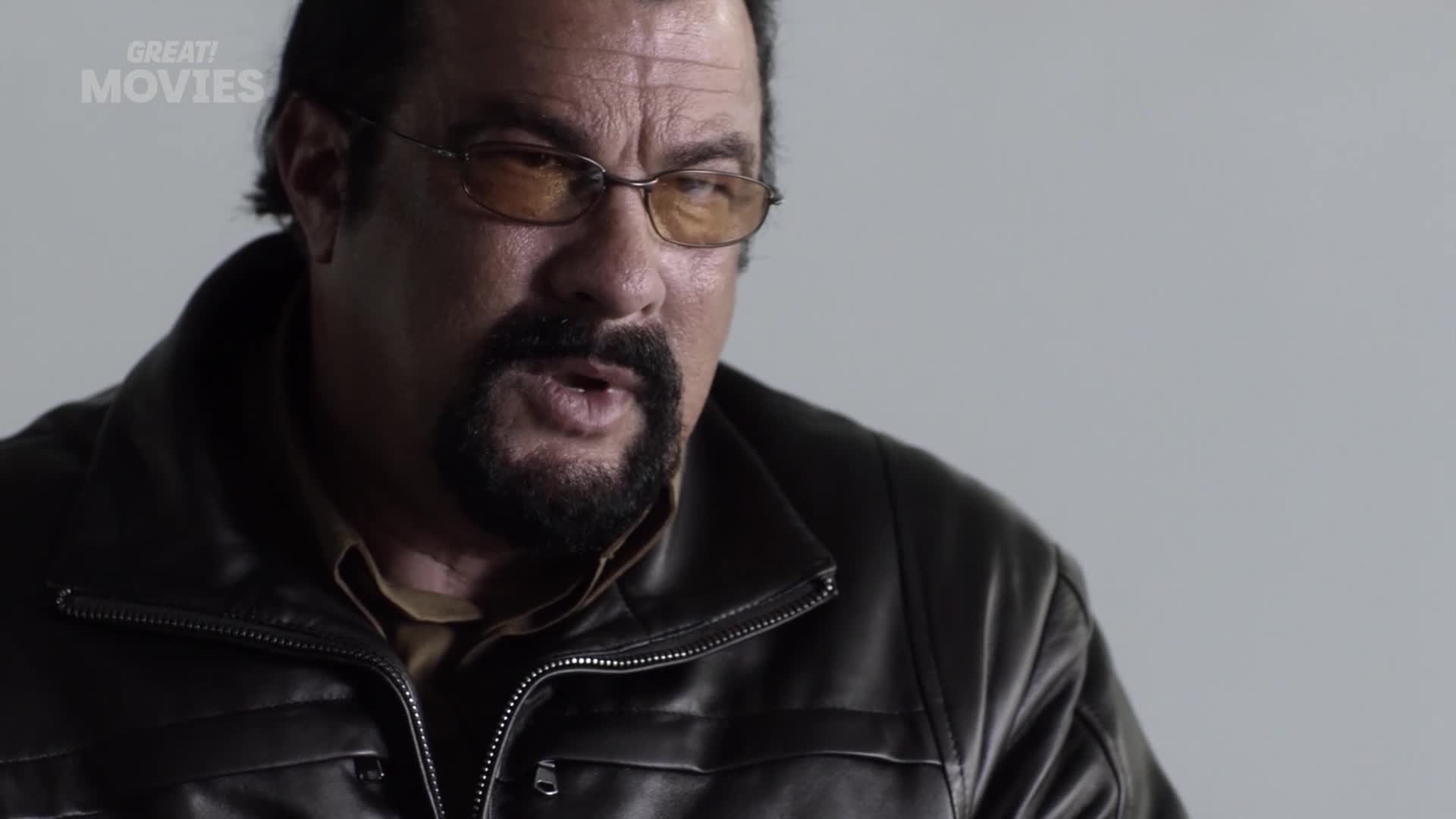 Steven Seagal, sporting his signature goatee and tinted glasses, speaks directly to the camera. He's wearing a dark leather jacket over a collared shirt, looking every bit the action star you'd expect from a GREAT! Movies broadcast.