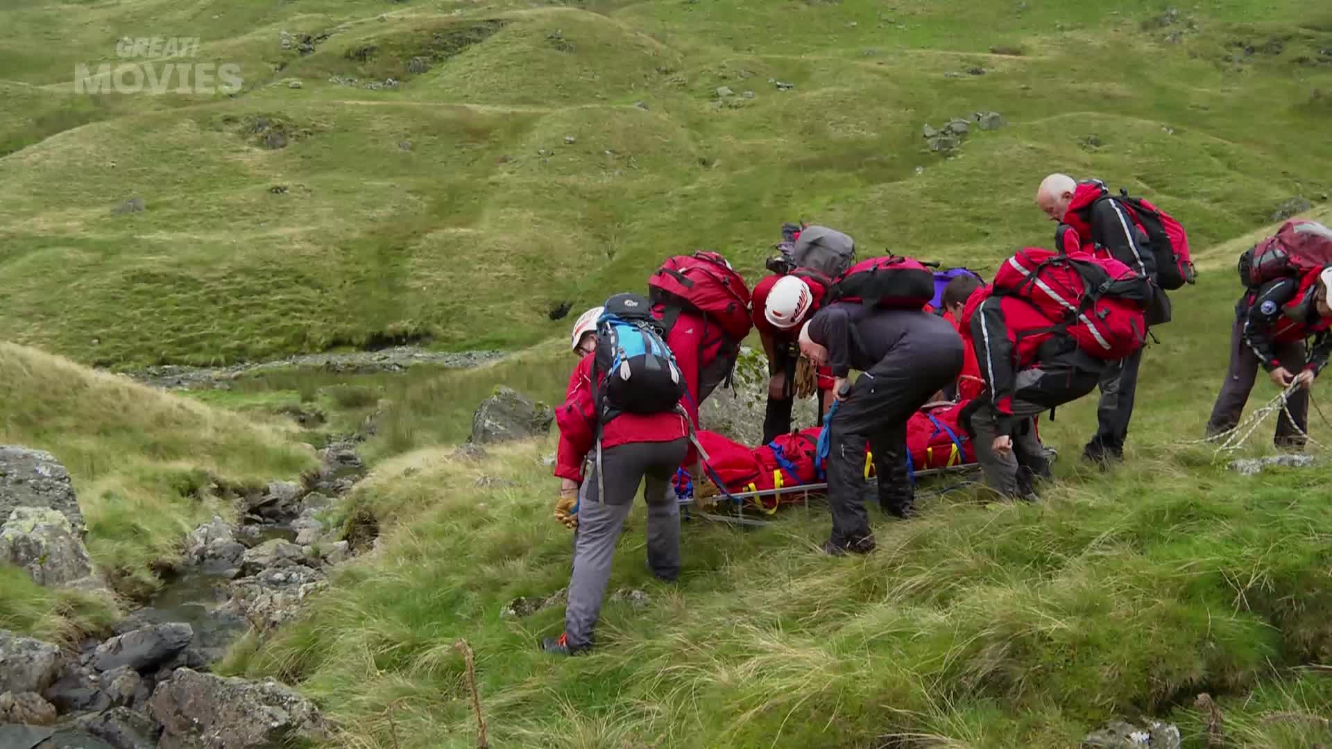 A team of rescuers in bright red jackets are carefully maneuvering a stretcher down a grassy, uneven slope. They're working together, their large backpacks a common sight in the British countryside.