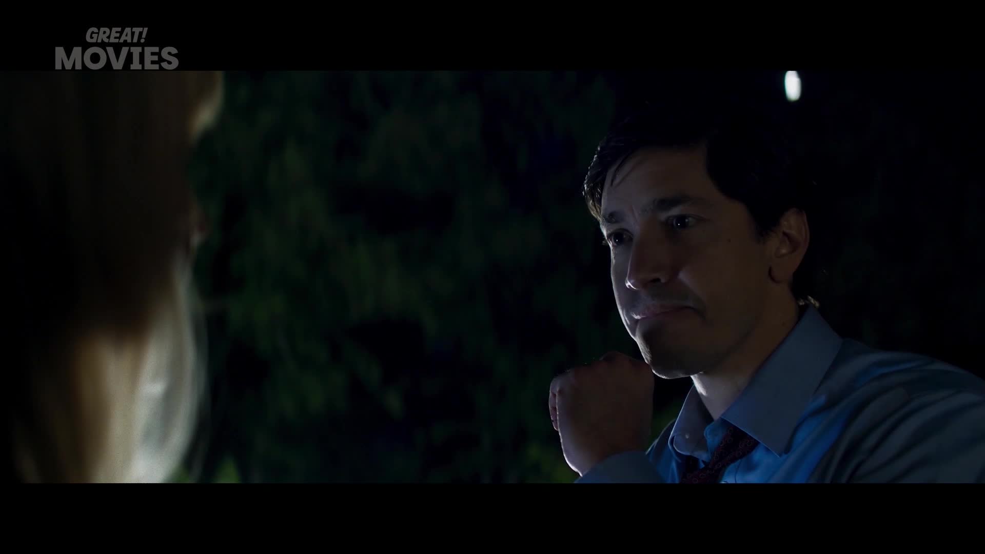 A man in a blue shirt and tie stares intently forward, his brow furrowed. The scene unfolds at night, with dark trees forming a backdrop.