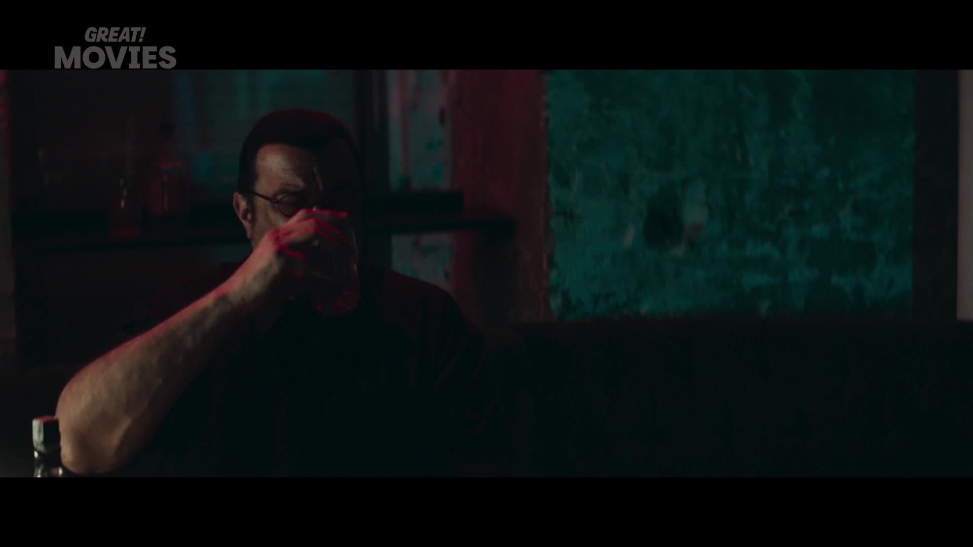 A man with glasses, his face partially obscured by shadow, lifts a glass to his lips. The dim lighting, tinged with red and green, suggests a late-night scene in a London pub.