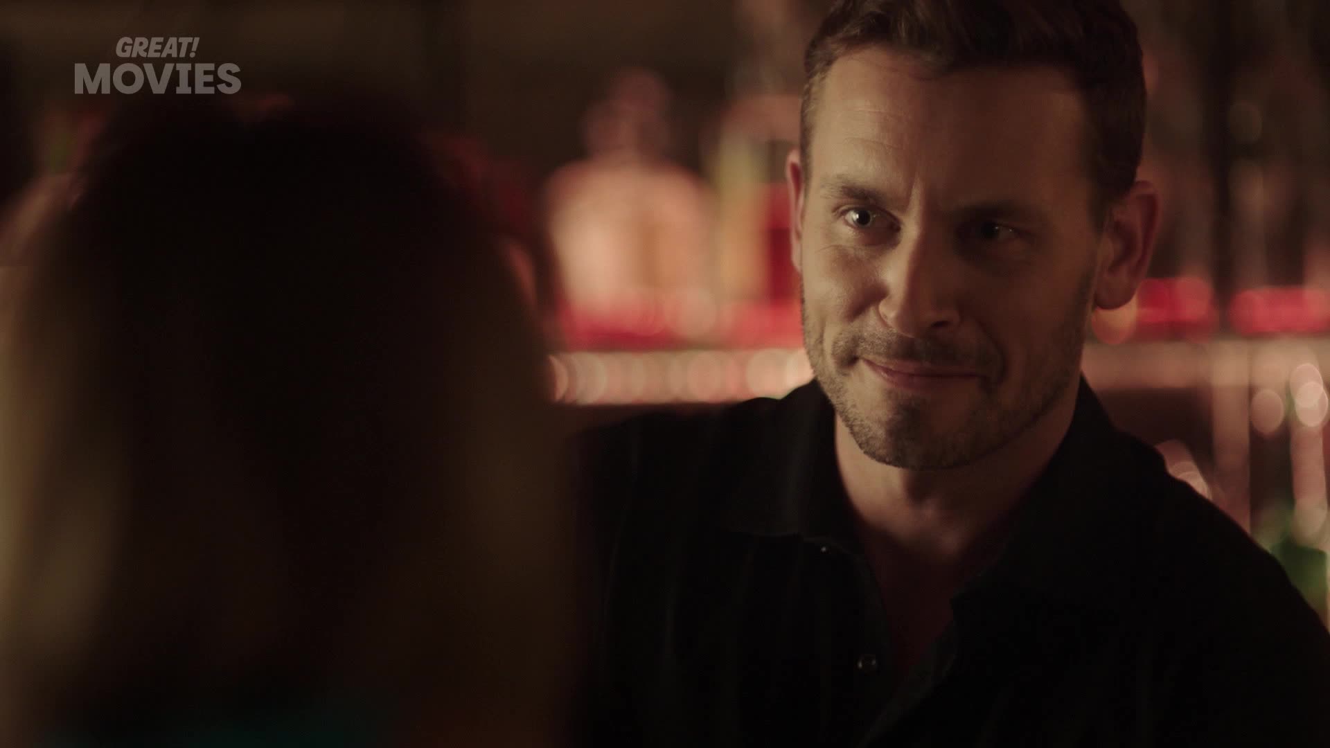 A man with a slight smile leans in, his eyes fixed on someone just out of frame. Behind him, the warm glow of a bar lights up bottles on shelves.