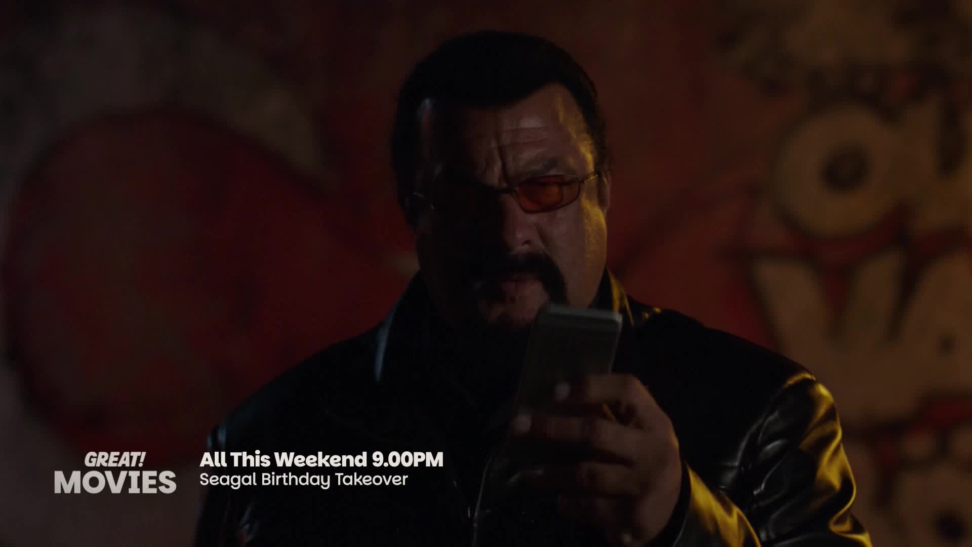 Steven Seagal stands in shadow, holding a phone. A red mural is visible behind him.