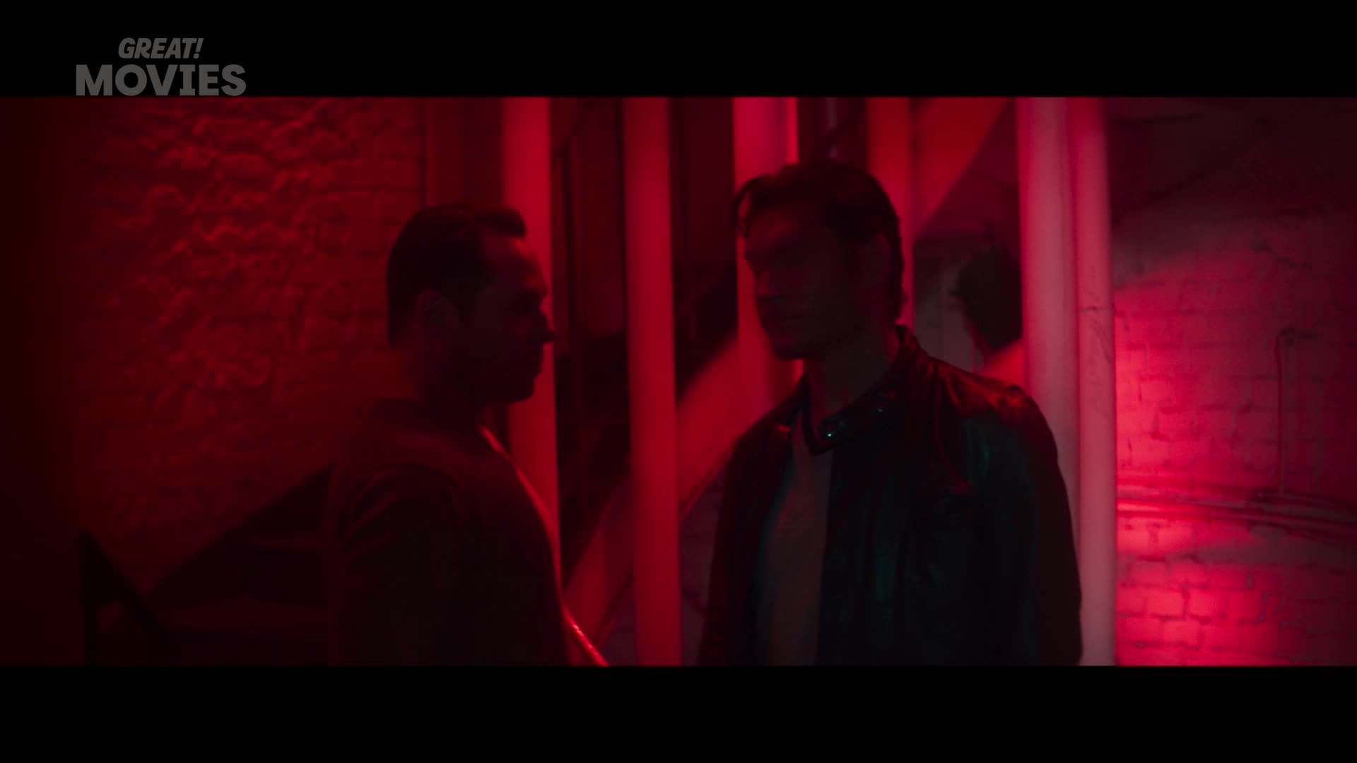 Two men stand in a dimly lit space, bathed in a strong red light. The man on the left, facing away, appears to be speaking.