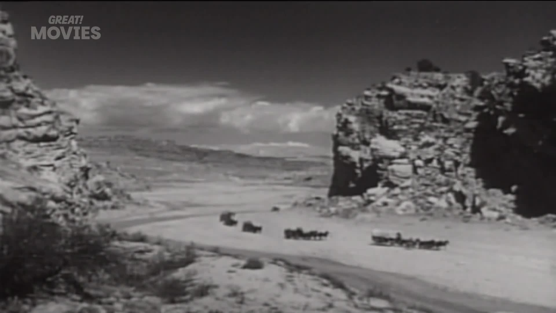 A line of horse-drawn wagons kicks up dust as they travel along a dirt road. The wagons are making their way through a wide canyon with steep, rocky walls.
