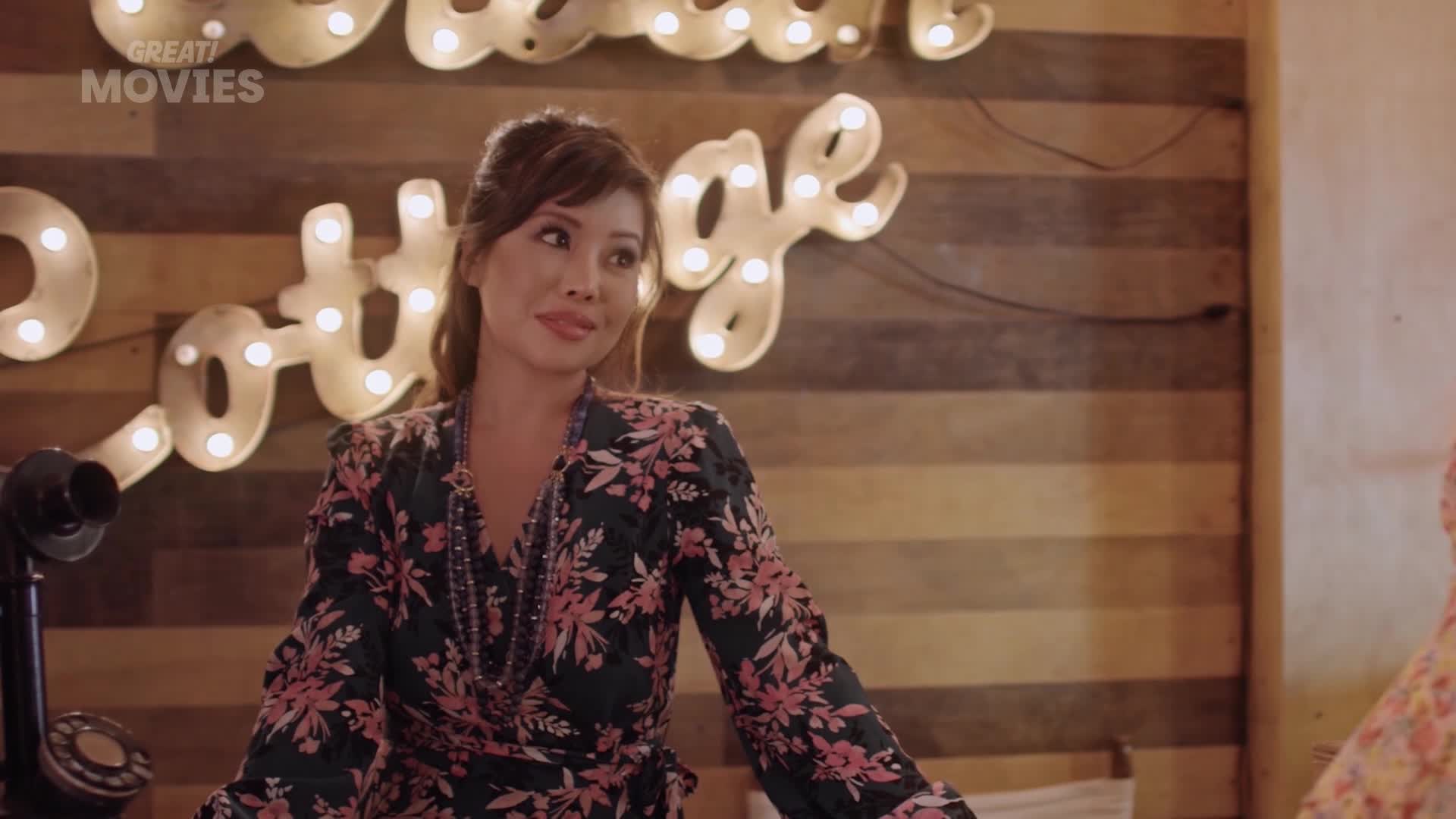 A woman in a floral dress stands by a vintage telephone. Behind her, illuminated letters spell out "Cottage" against a wooden wall.
