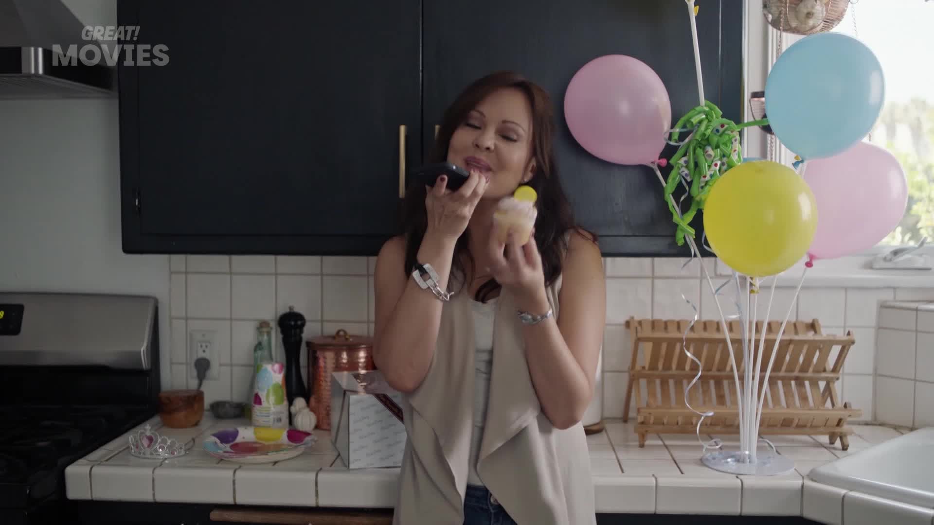 A woman holds a cupcake to her mouth while speaking into a phone. Pastel balloons bob on strings behind her, a festive touch for any GREAT! movie moment.