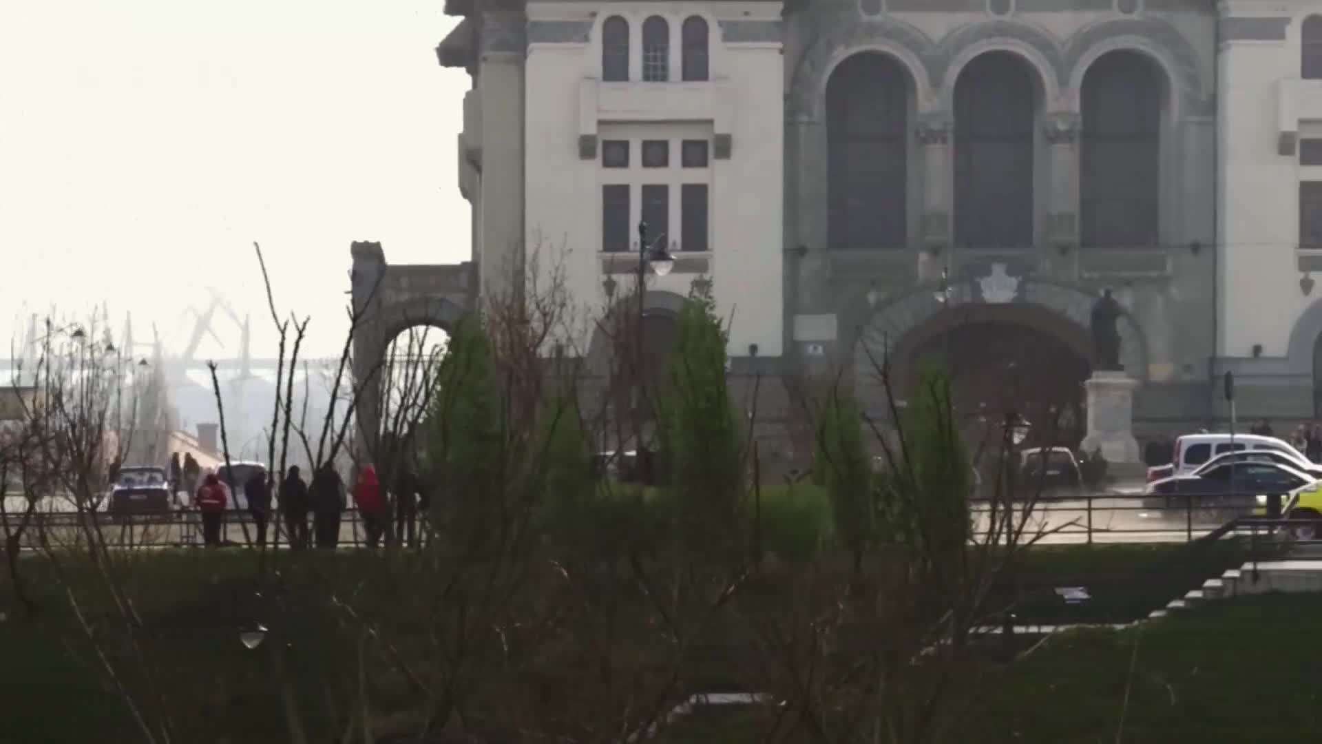 A line of people strolls past a dark car on the pavement. A white van and a yellow car pass by the grand building.