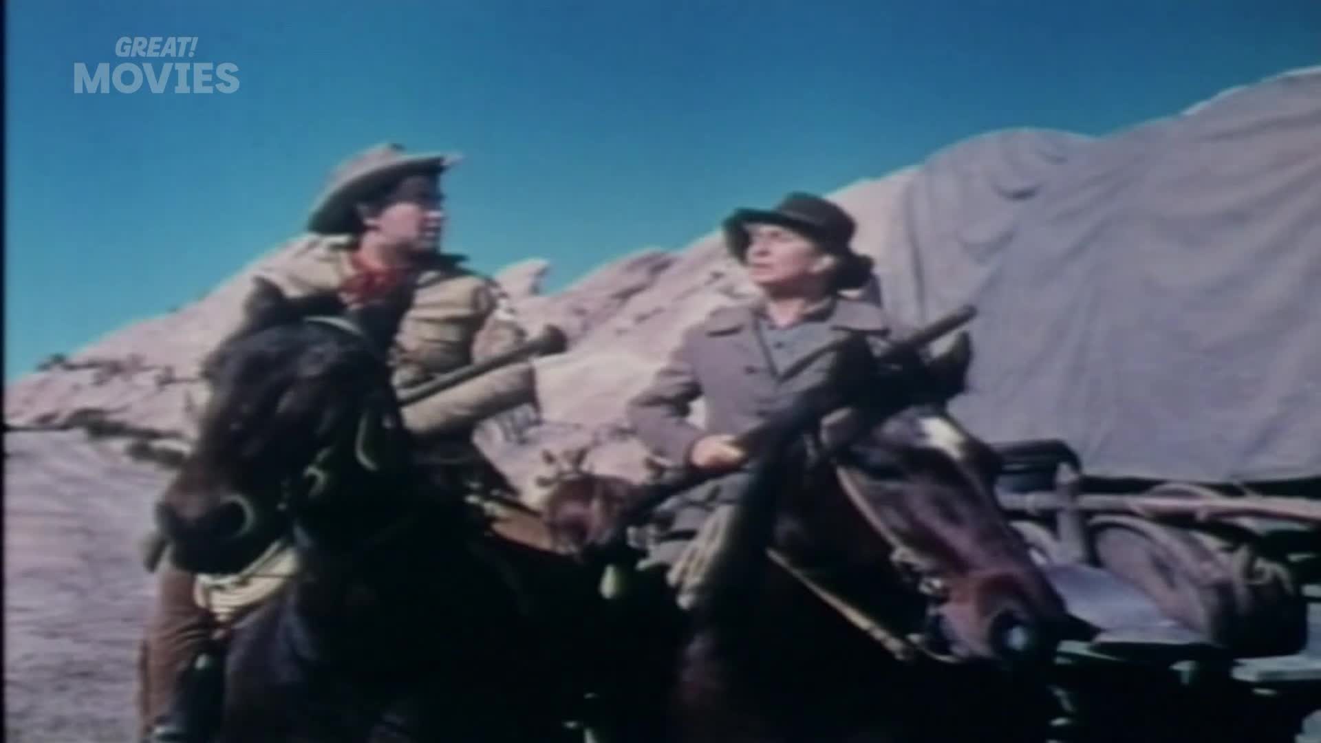 Two riders on horseback, a man in a cowboy hat and a woman in a brimmed hat, look towards a covered wagon. The horses stand patiently against a backdrop of rocky hills and a bright blue sky.
