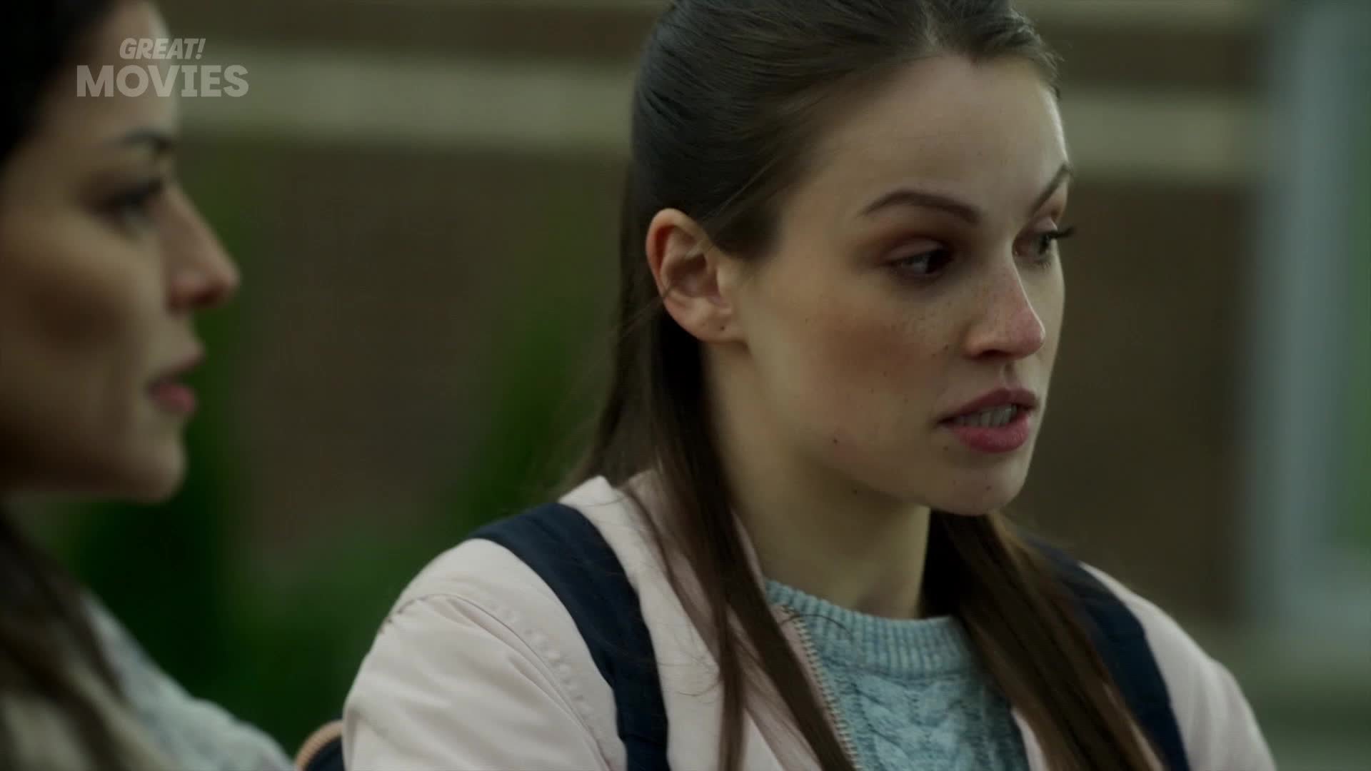 Two young women are talking, their faces turned towards each other. The one on the right, with freckles across her nose, speaks with a slight frown.