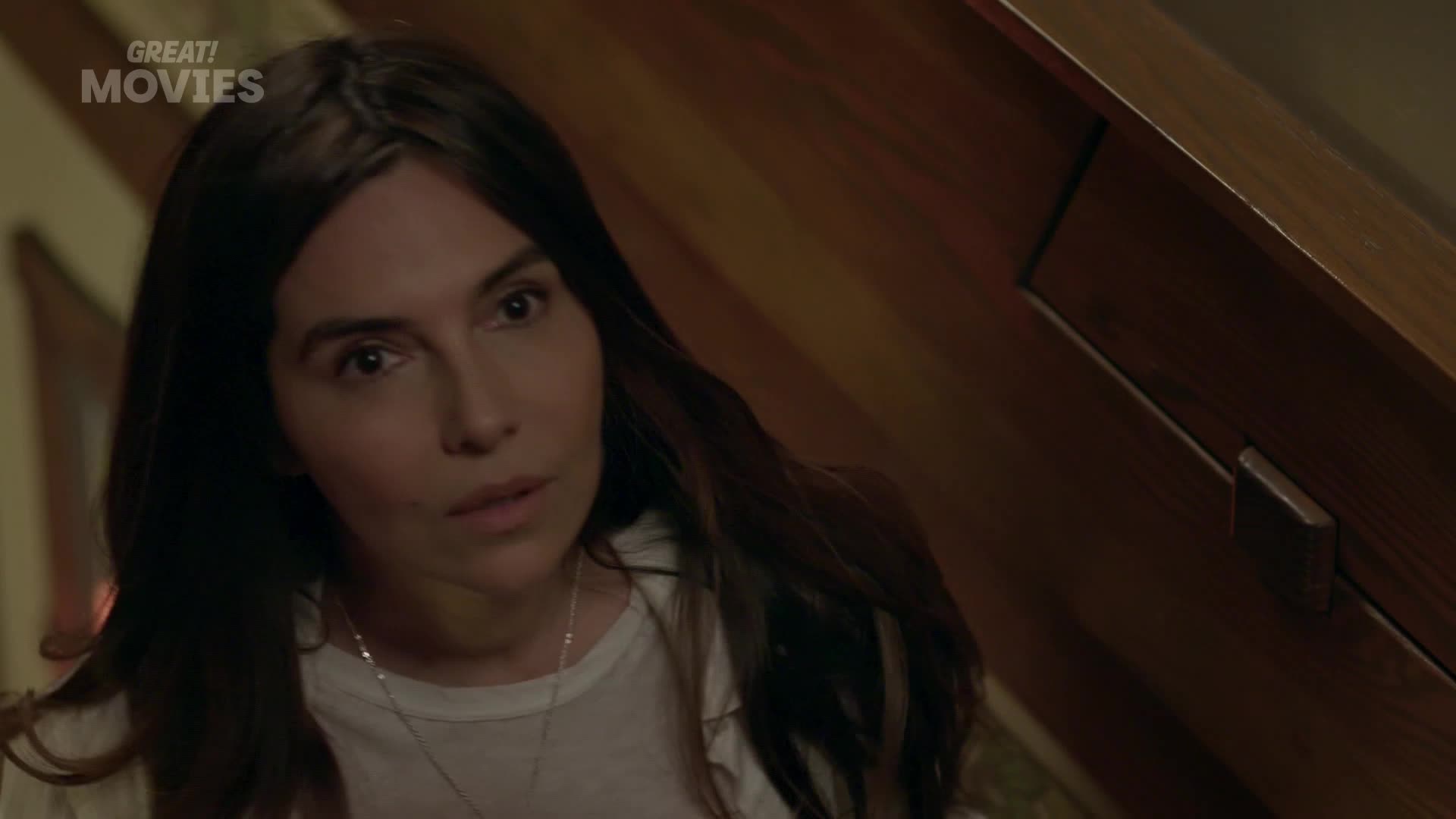 A woman with long, dark hair turns her head, her expression a mixture of concern and curiosity. The warm glow of a lamp illuminates the wooden paneling behind her.