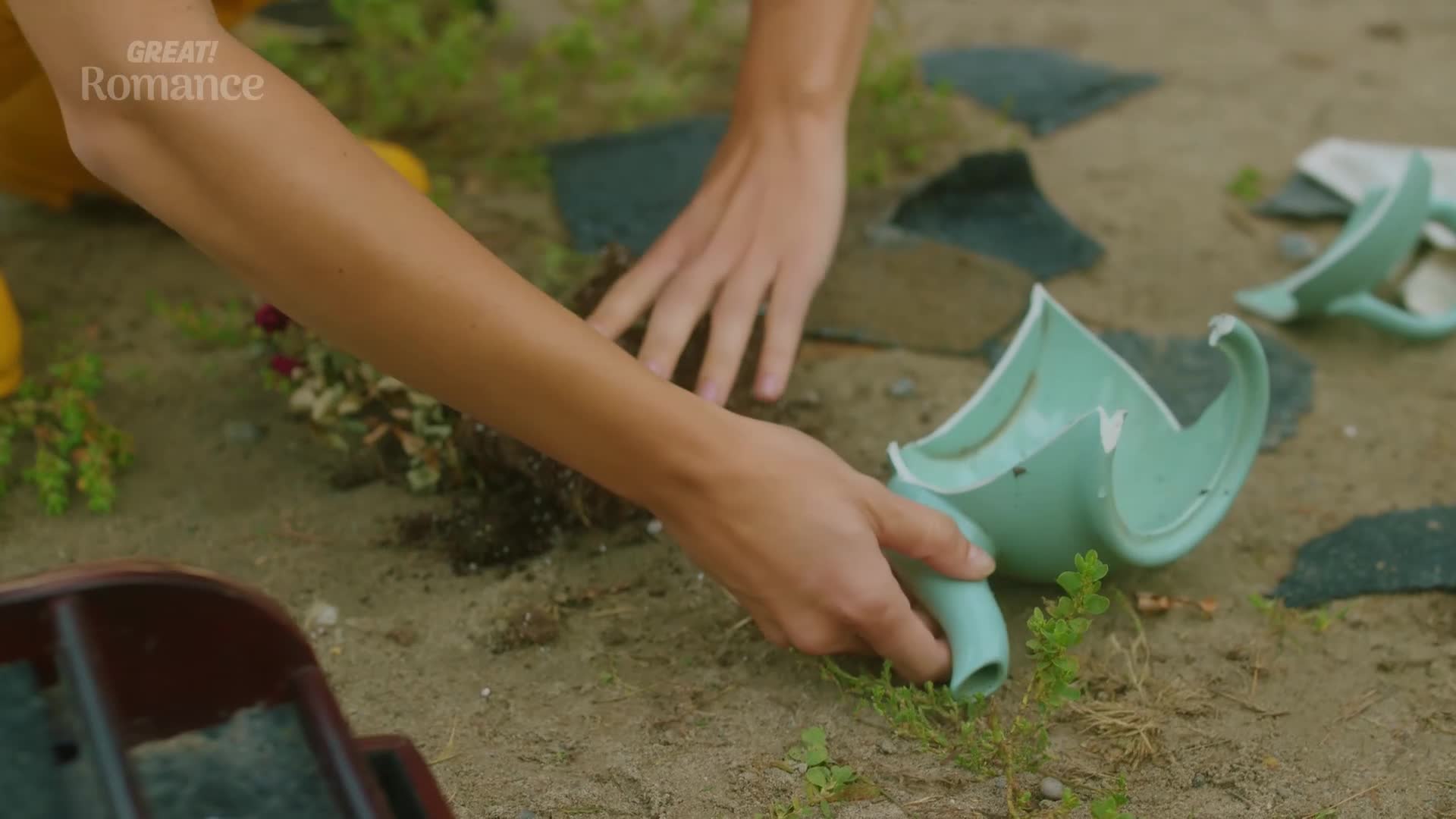 Hands sift through dirt, gathering up pieces of a broken pale green watering can. Scattered around are shards of the same ceramic, some sharp, some smooth, lying on the sandy ground.