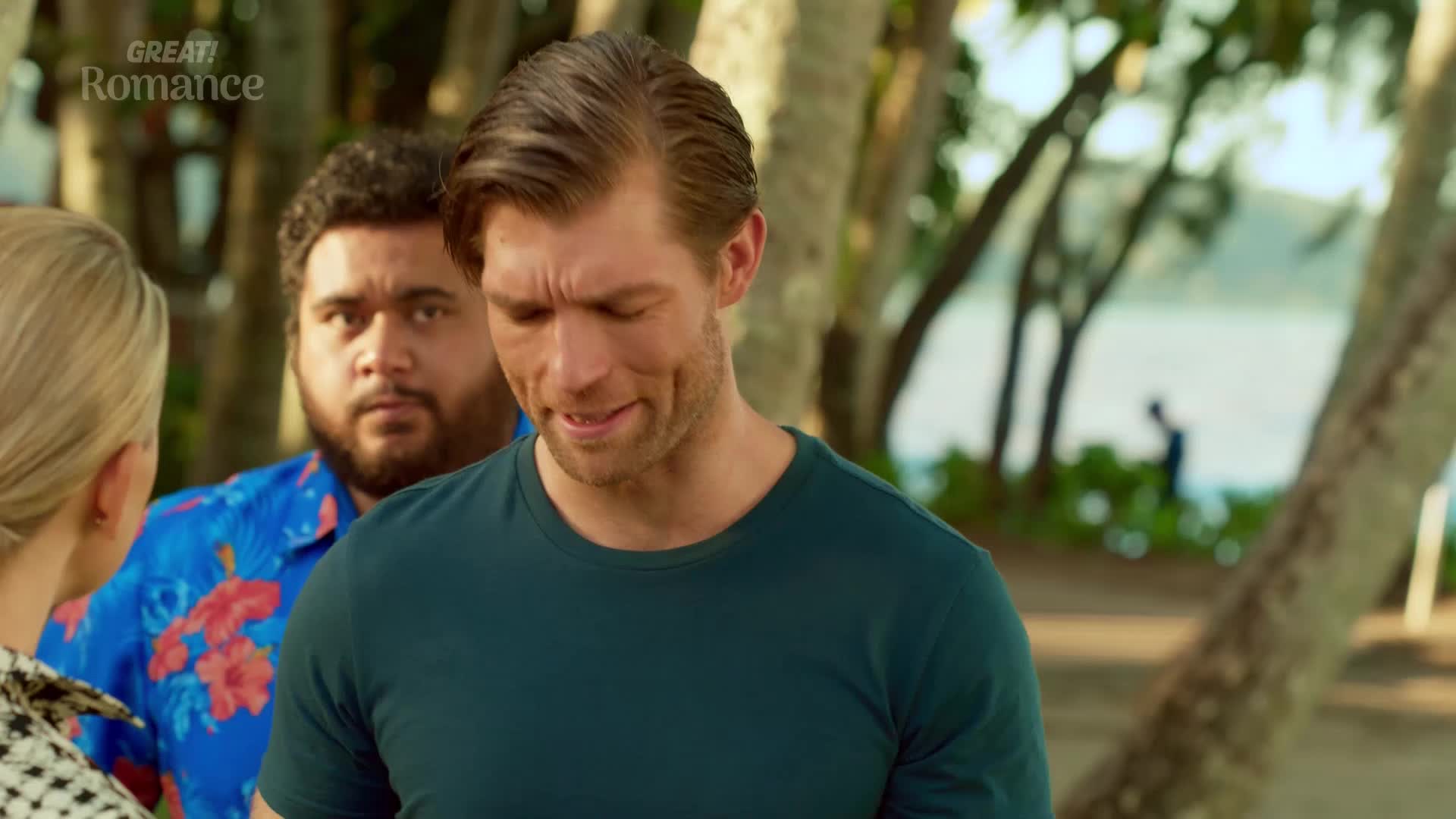 A man in a teal shirt looks down, his brow furrowed. Behind him, another man in a bright floral shirt watches intently.