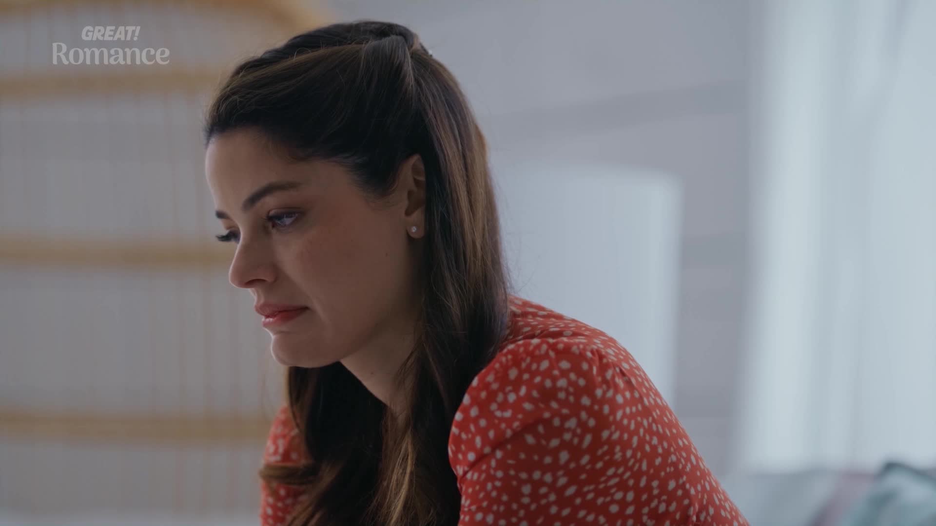 A woman with dark hair, wearing a red dress, looks down with a pensive expression. The faint "GREAT! Romance" logo suggests a scene from a British film.