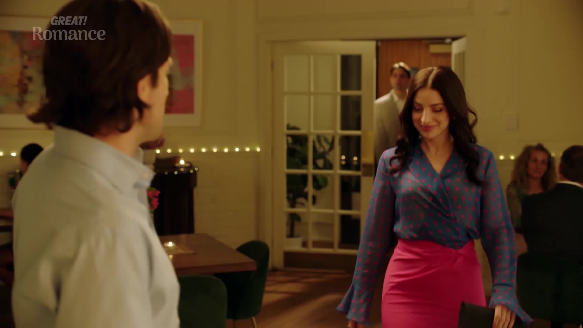 A man in a pale blue shirt turns his head as a woman in a vibrant pink skirt and patterned blouse walks past. She glances down, a slight smile playing on her lips, as a man in a light suit stands in the doorway behind her.