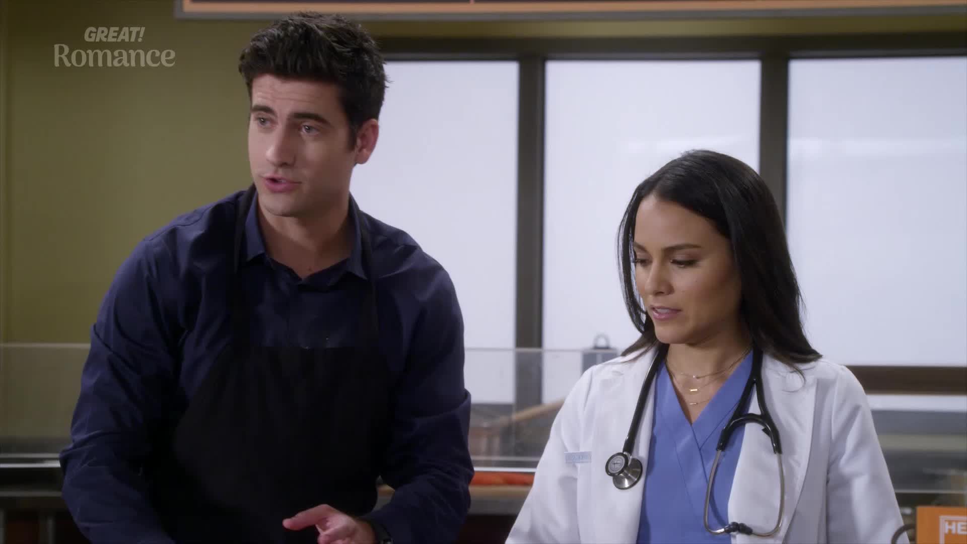 A man in a dark blue shirt and apron gestures as he speaks to a woman in a white doctor's coat. She listens intently, her stethoscope resting around her neck.