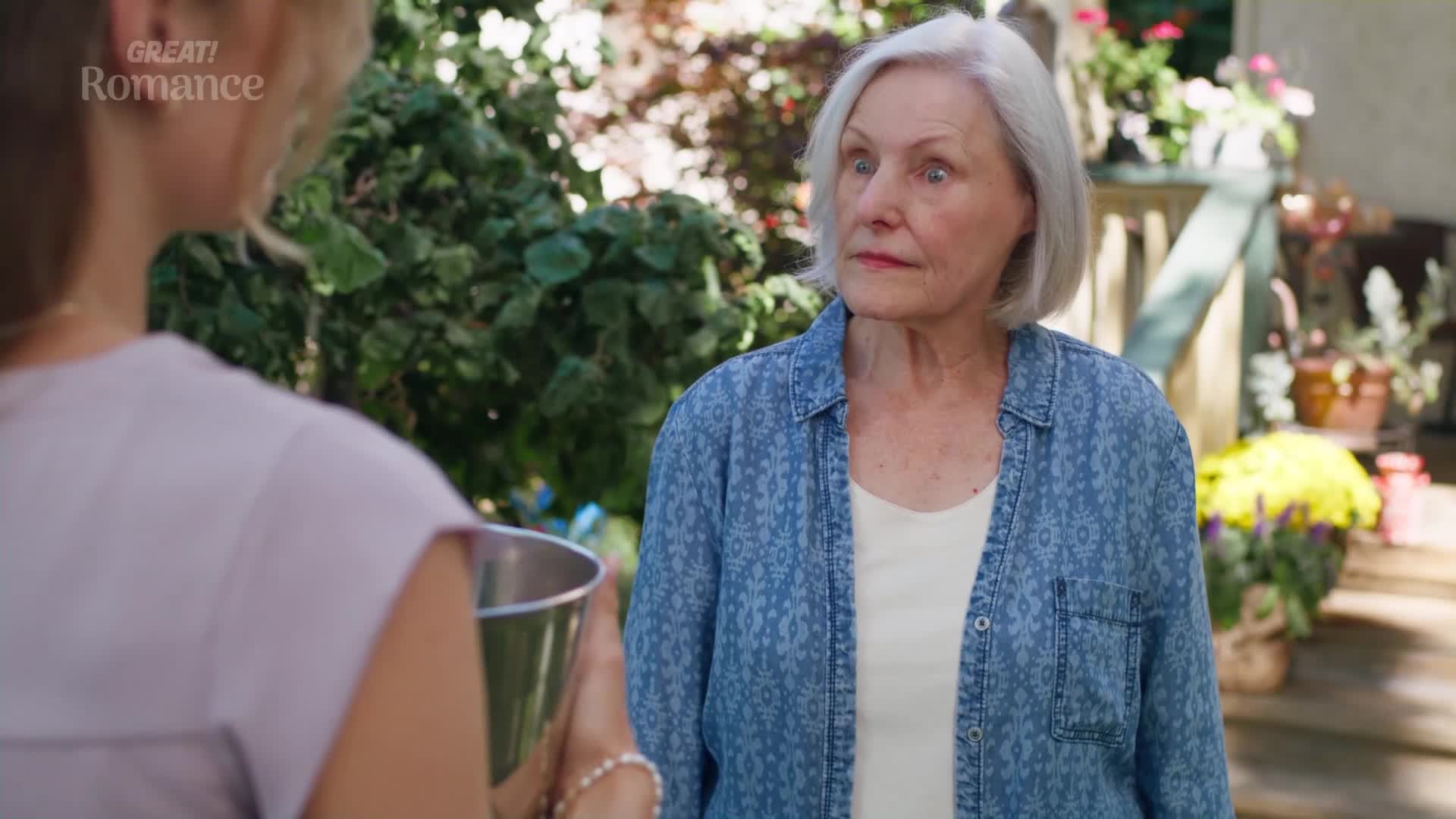 The older woman's eyes widen as the younger woman approaches with a metal bucket. Sunlight filters through the leaves of the garden behind them.