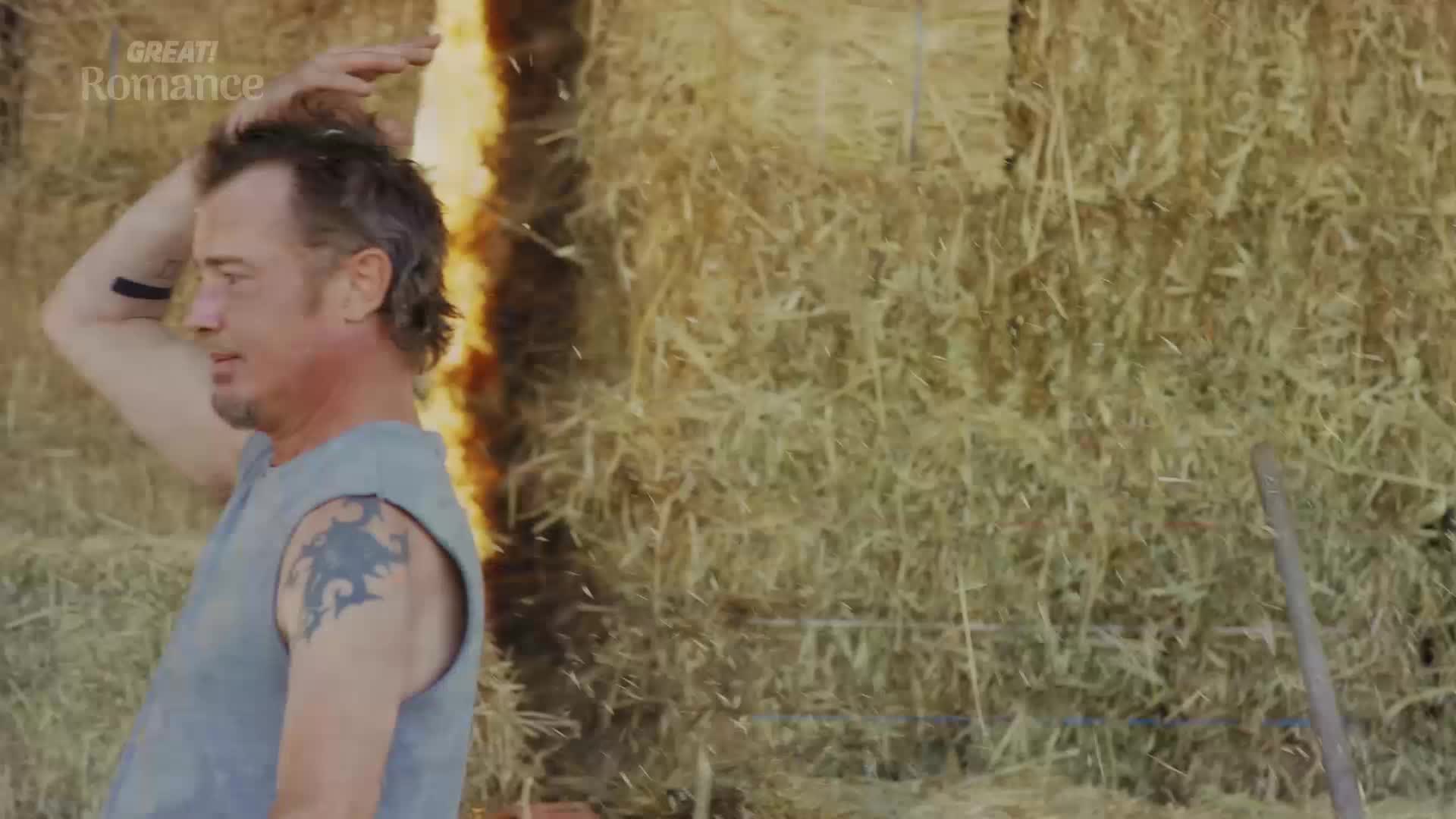A man with a tribal tattoo on his arm scratches his head, standing beside a wall of hay bales. A bright orange flame licks up the side of the bales, casting a warm glow.