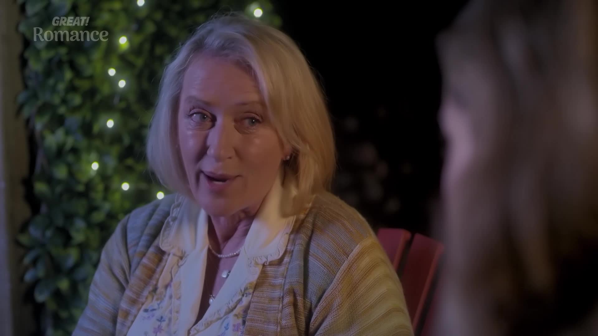 A woman with blonde hair, wearing a patterned cardigan, speaks animatedly. Twinkling lights are visible behind her.