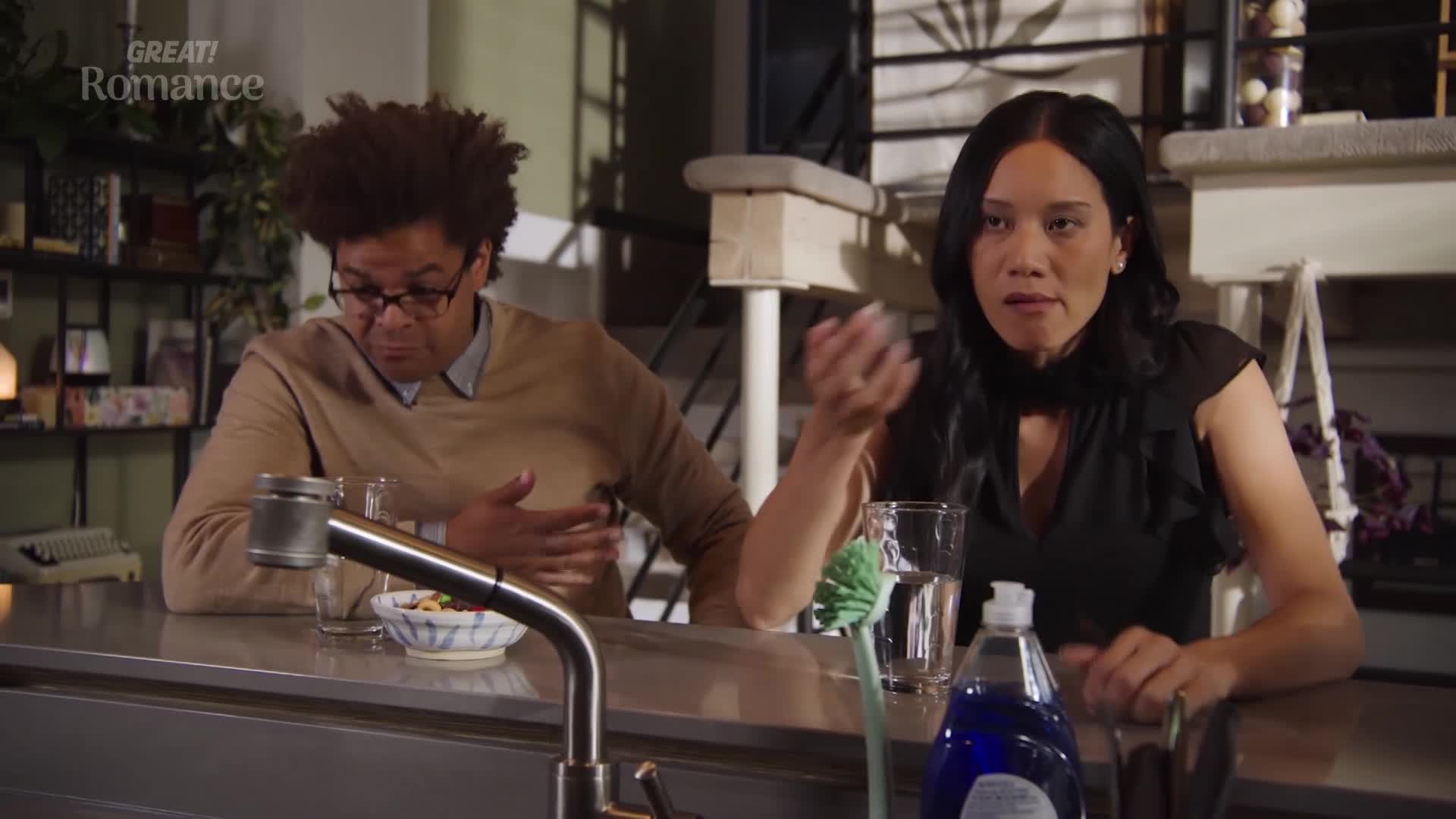 A woman with dark hair gestures emphatically as she speaks, her brow furrowed. Beside her, a man with glasses and a brown sweater leans forward, his expression thoughtful.