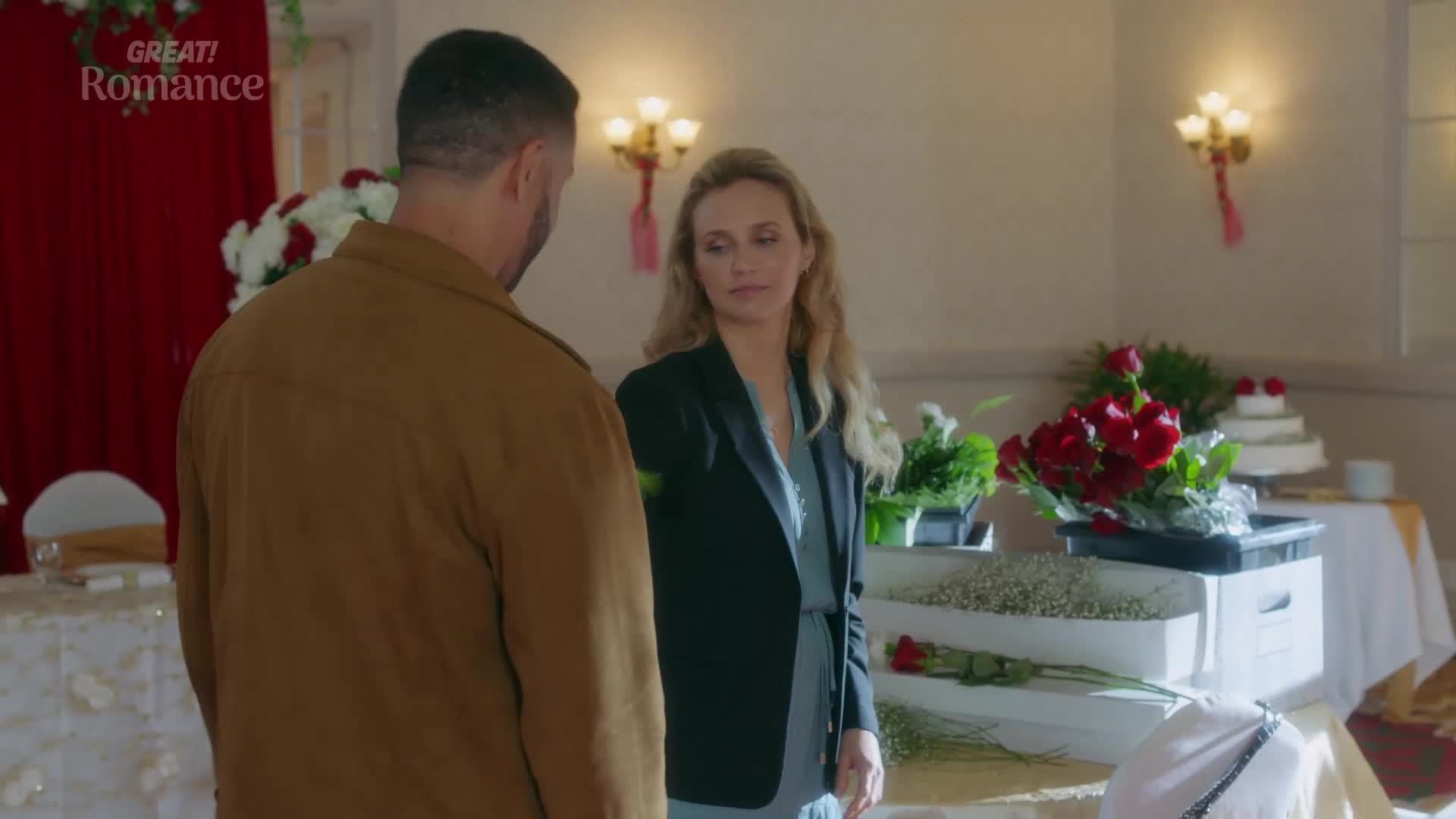 A man in a tan jacket stands with his back to me, facing a woman in a dark blazer. Behind them, a table is laden with white flowers and a large bouquet of red roses, with a tiered cake visible on a side table.