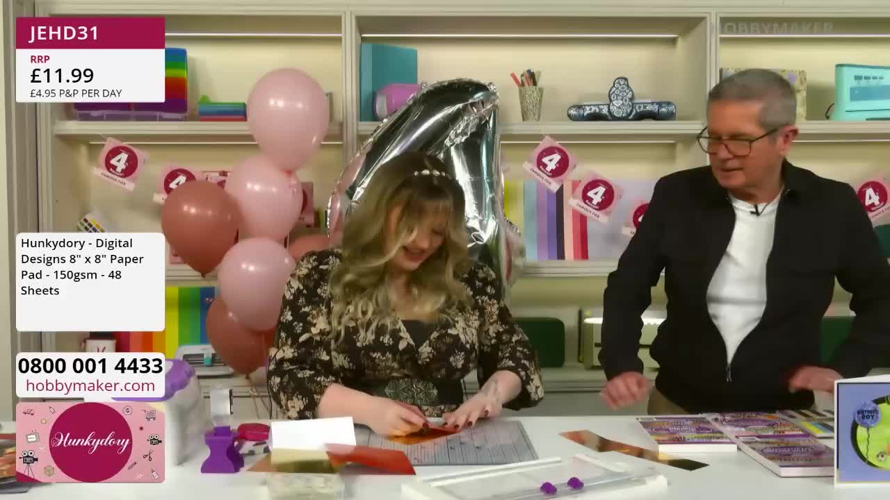 A woman is carefully cutting a piece of paper on a mat, while a man watches her with a smile. Pink balloons float behind them, and shelves filled with craft supplies are visible in the background. A woman is carefully cutting a piece of paper on a mat, while a man watches her with a smile. Pink balloons float behind them, and shelves filled with craft supplies are visible in the background.