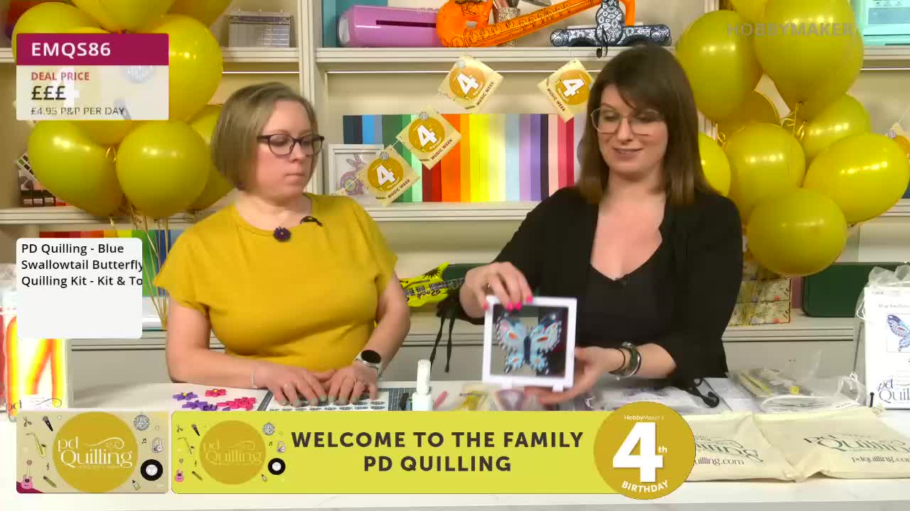 A woman in a black cardigan holds up a framed quilled butterfly. Beside her, a woman in a yellow top looks down at a crafting surface. Yellow balloons cluster behind them. A woman in a black cardigan holds up a framed quilled butterfly. Beside her, a woman in a yellow top looks down at a crafting surface. Yellow balloons cluster behind them.