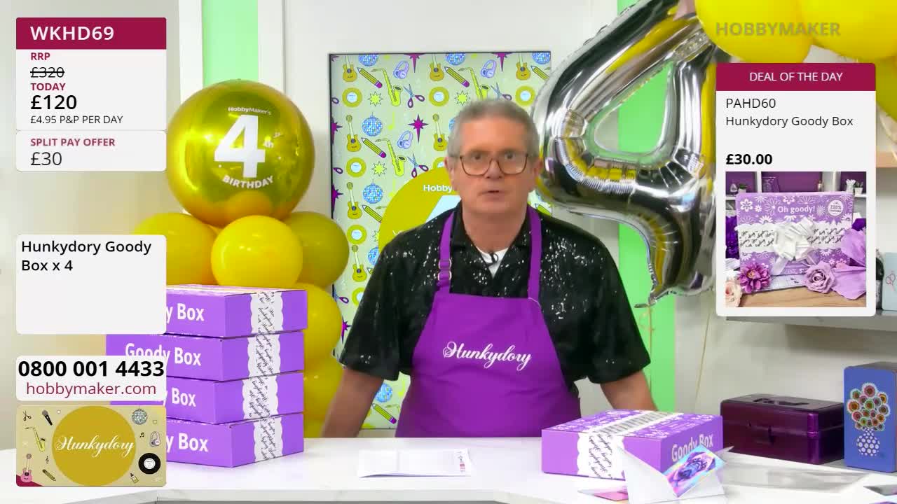 A man in a sparkly shirt and a purple apron stands behind a table laden with purple boxes. A giant silver "4" balloon looms behind him, next to a cluster of yellow balloons. A man in a sparkly shirt and a purple apron stands behind a table laden with purple boxes. A giant silver "4" balloon looms behind him, next to a cluster of yellow balloons.