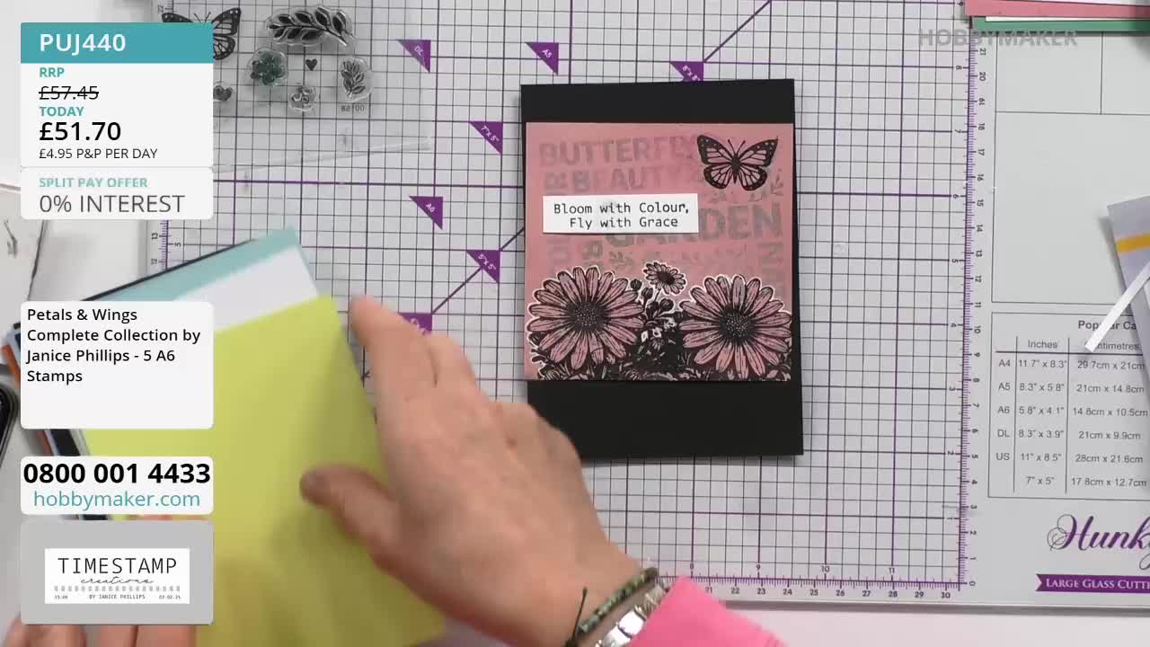 A hand slides a bright yellow cardstock behind a finished card featuring daisies and a butterfly. The card, a Hobby Maker product, is displayed on a cutting mat.